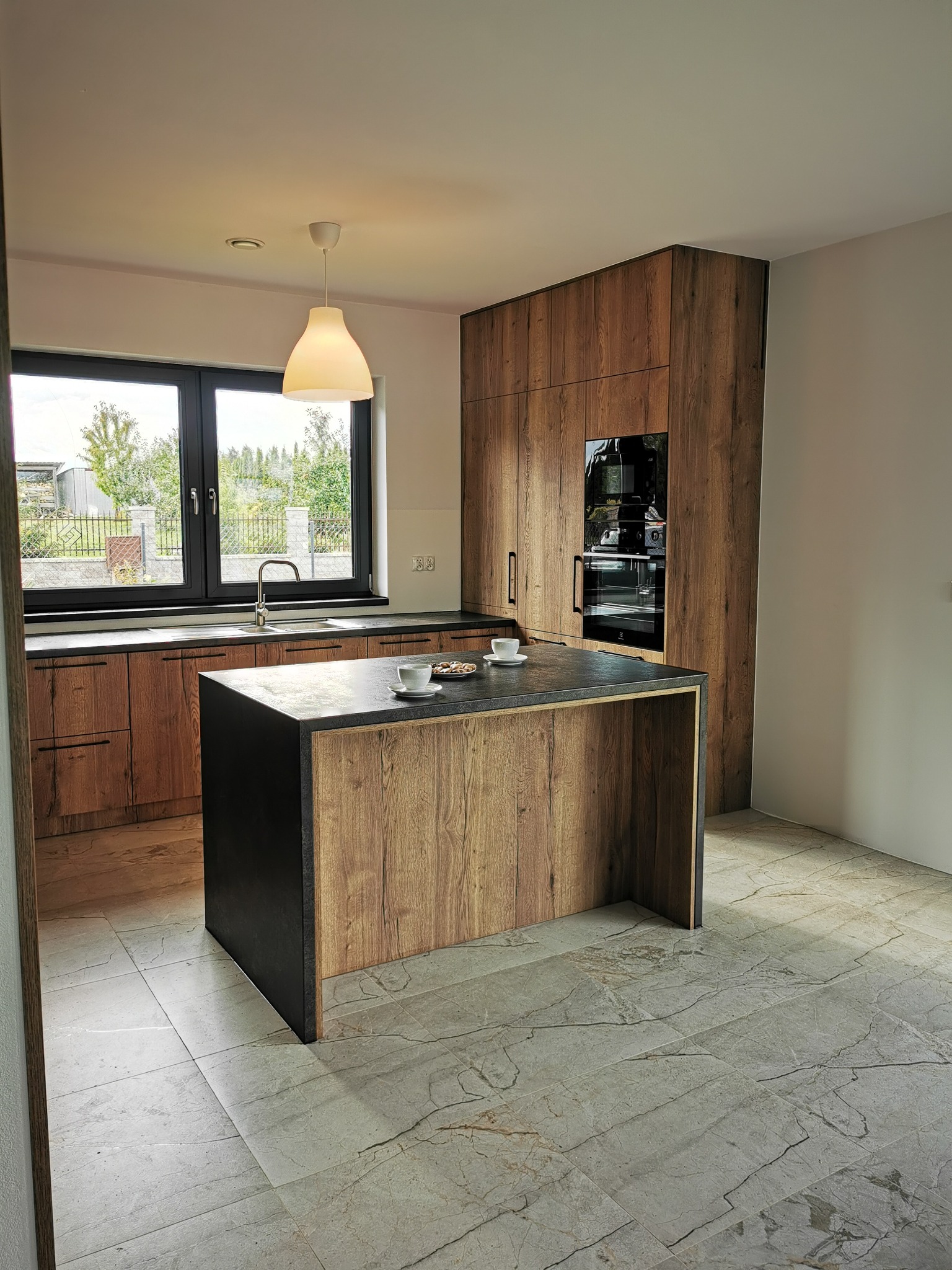 Nowoczesna kuchnia z drewnianymi frontami, wyspą z czarnym blatem i dwoma filiżankami, widok przez okno na zieleń.