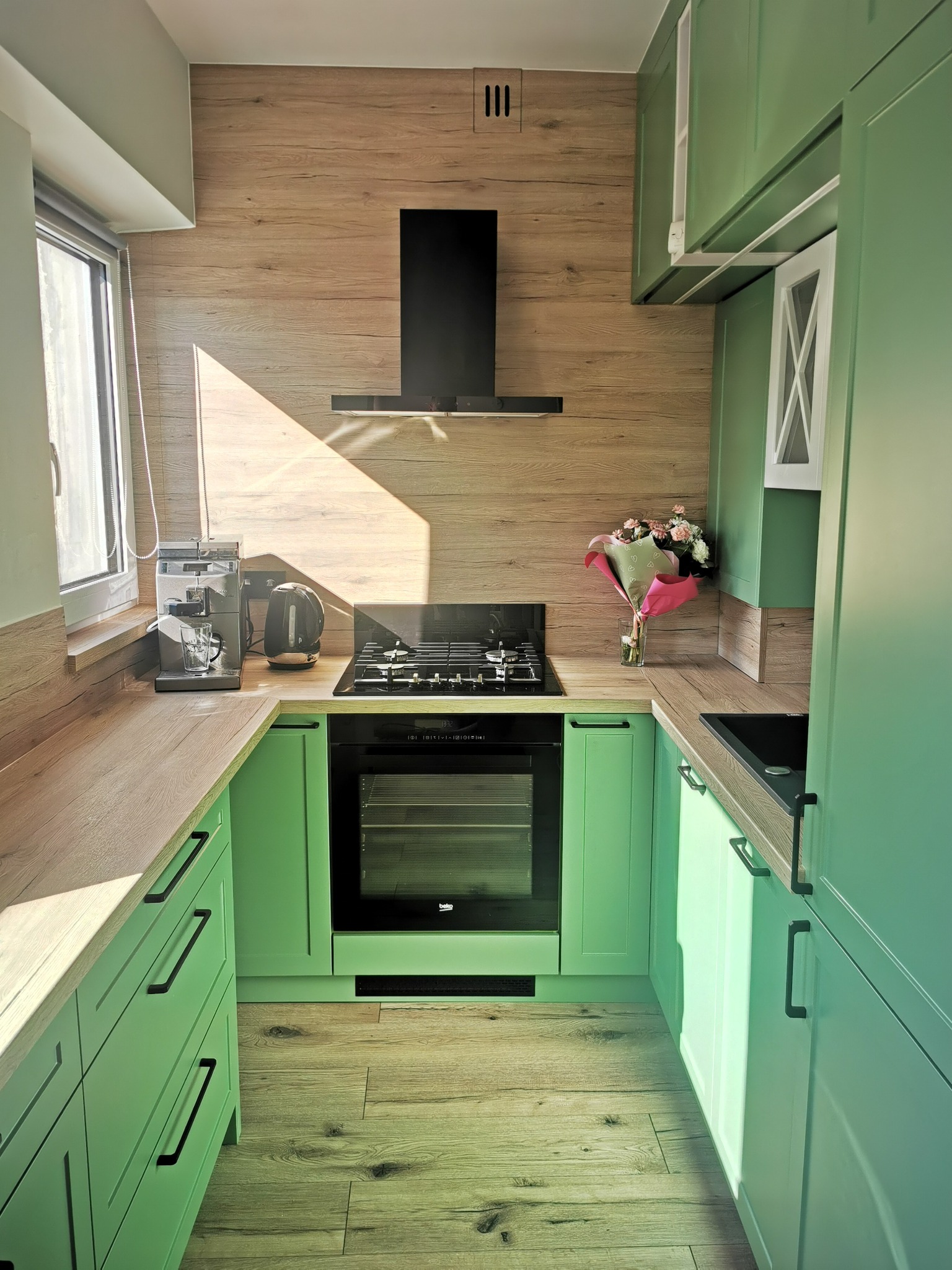 Jasnozielona kuchnia z drewnianymi akcentami w Motyczu, wyposażona w czarny okap i płytę gazową, oświetlona promieniami słońca wpadającymi przez okno.