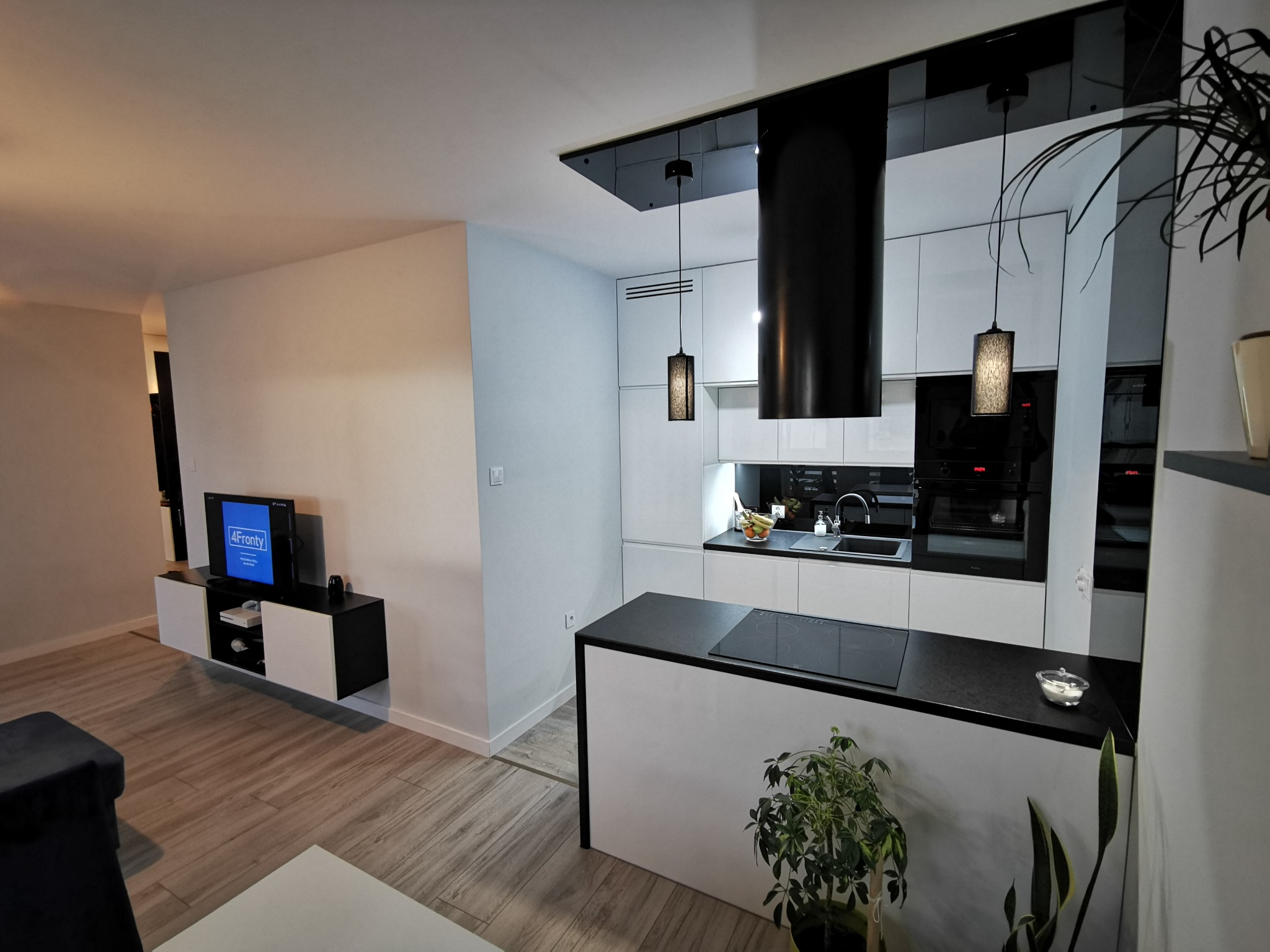 Nowoczesna kuchnia z białymi frontami, czarnym blatem i wyspą, oświetlona wiszącymi lampami z elementami czerni, widok na salon z telewizorem.