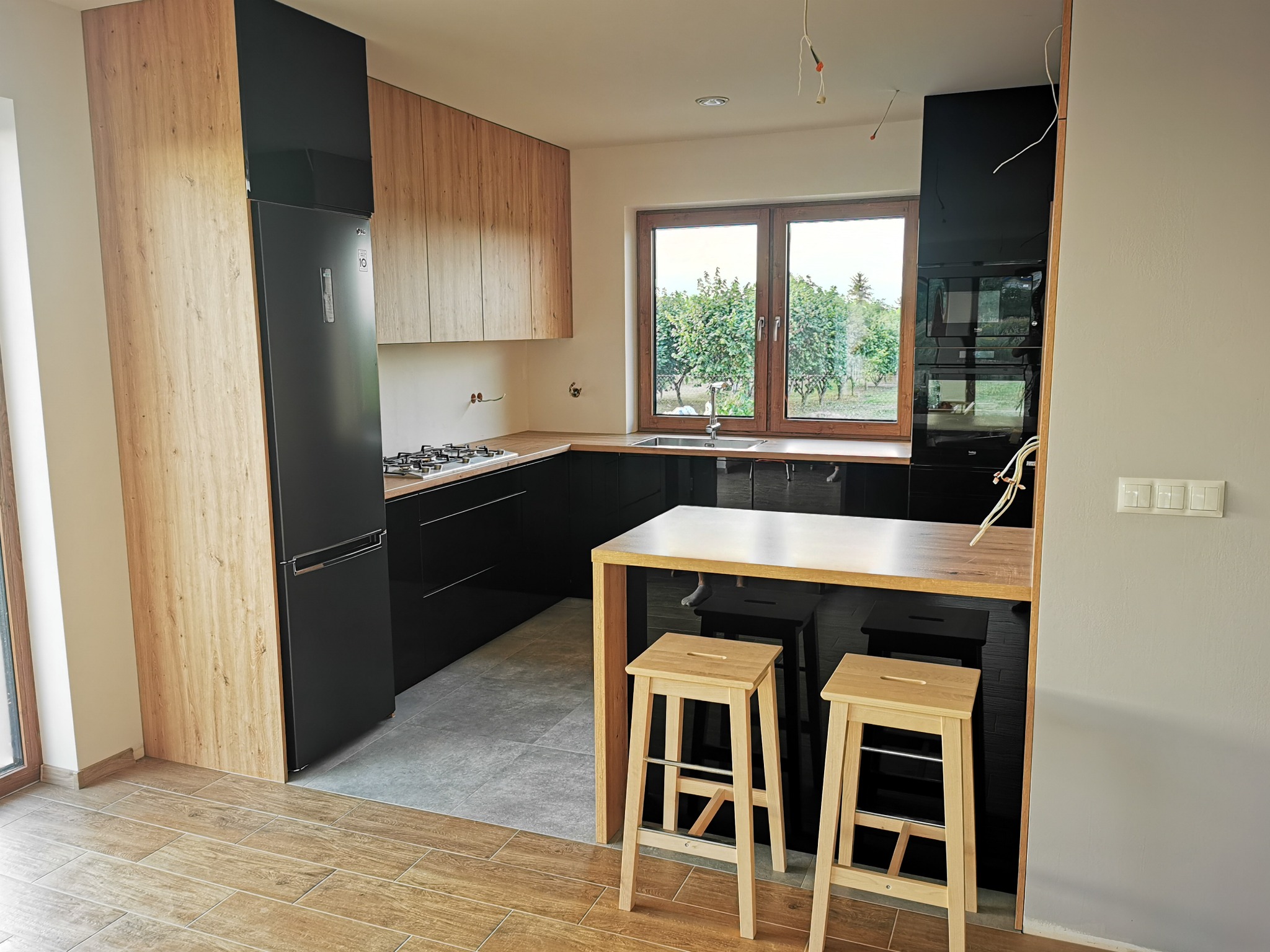 Nowoczesna kuchnia z czarnymi szafkami dolnymi i drewnianymi górnymi, wyspa z dwoma drewnianymi stołkami, widok na ogród przez okno.