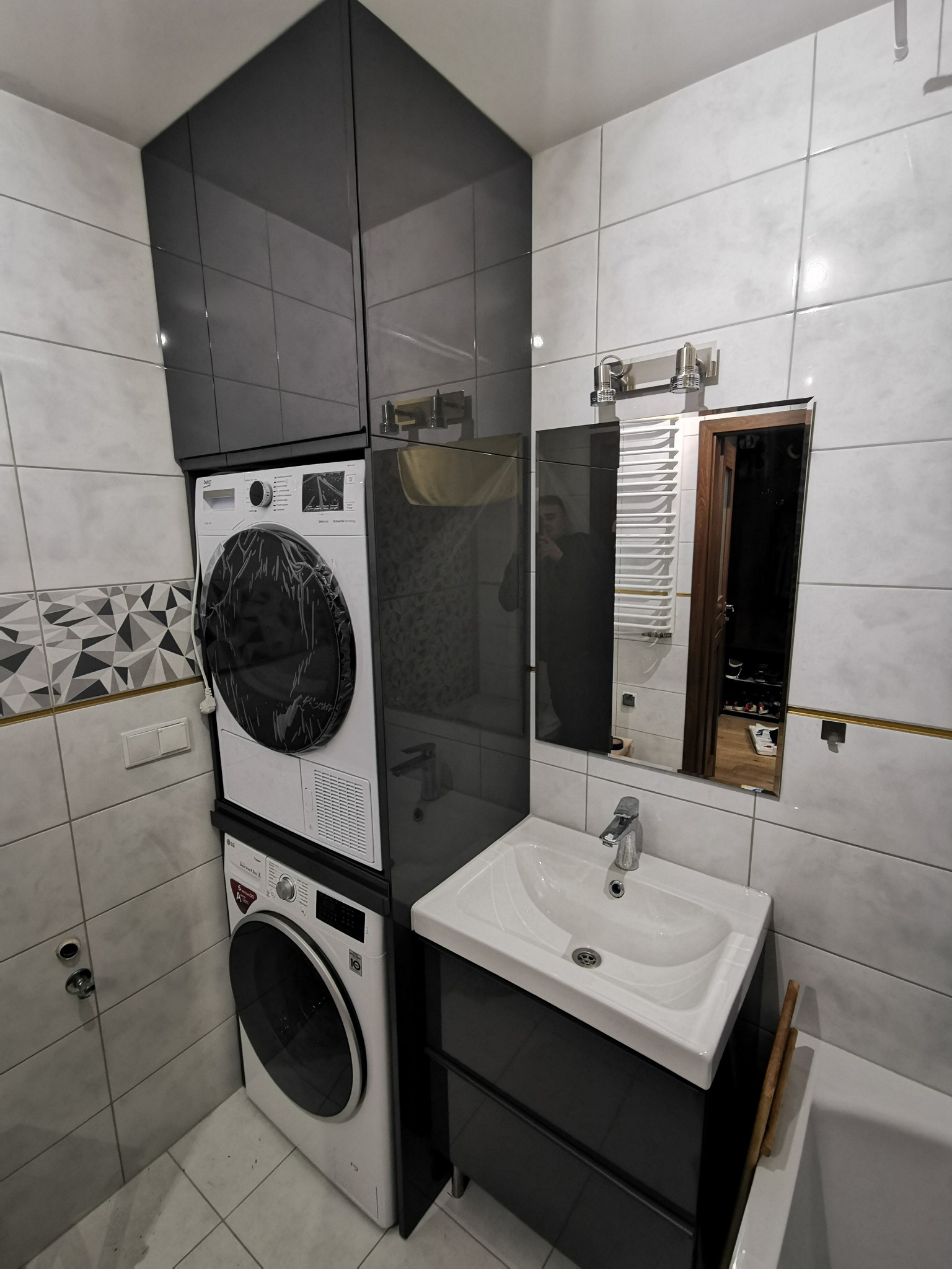 Ustawione pionowo pralka i suszarka w łazience, nad nimi szafki w kolorze antracytowym, obok umywalka z szafką w tym samym kolorze, całość na tle białych płytek.