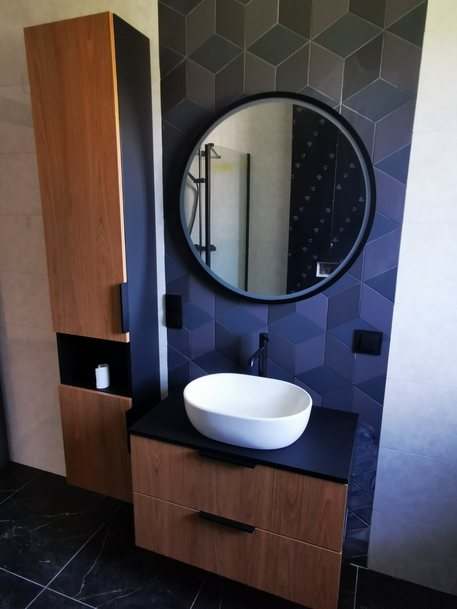 Nowoczesna łazienka z drewnianą szafką pod umywalką, okrągłym lustrem i geometrycznymi płytkami w odcieniach szarości i granatu.