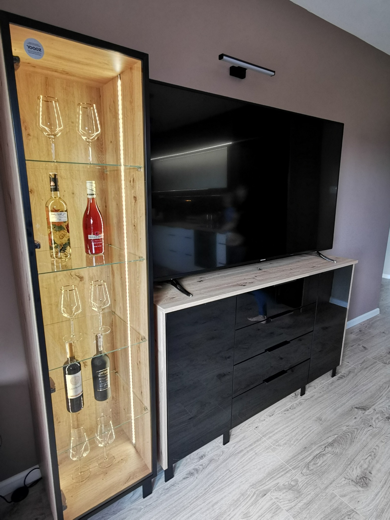 Nowoczesna szafka RTV z czarnymi frontami i blatem imitującym drewno, obok witryna z podświetleniem LED eksponująca kieliszki i butelki wina.