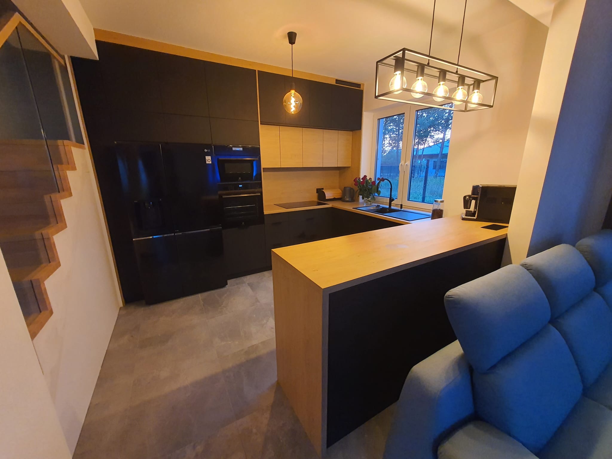 Nowoczesna kuchnia z czarnymi szafkami i drewnianymi akcentami, oświetlona lampą wiszącą z żarówkami Edison, z widokiem na zewnątrz przez okno i fragmentem schodów w tle.