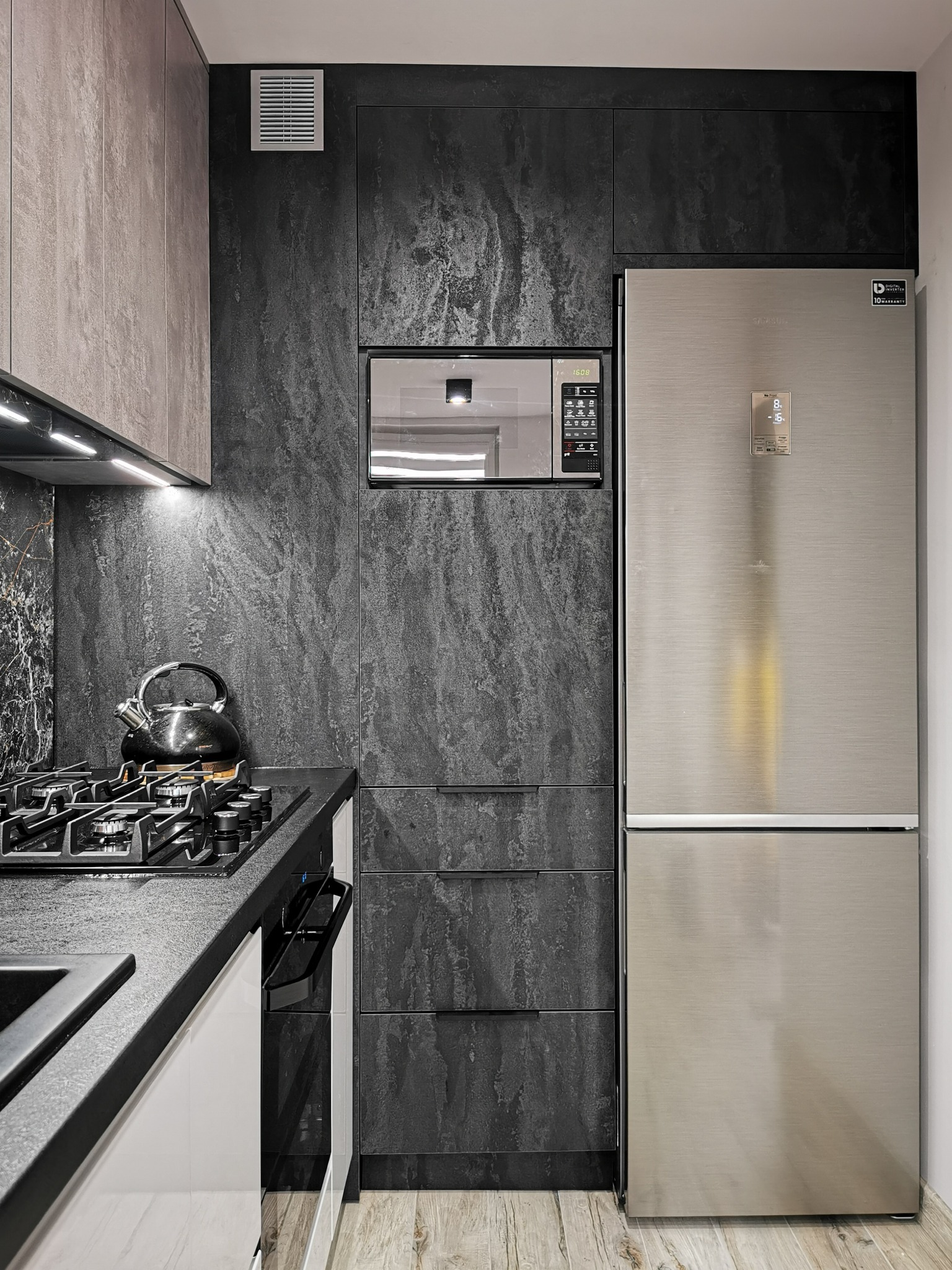 Częściowy widok nowoczesnej kuchni z czarnymi szafkami o teksturze kamienia, srebrną lodówką, czarnym blatem, płytą gazową z czajnikiem i białymi frontami szafek dolnych.