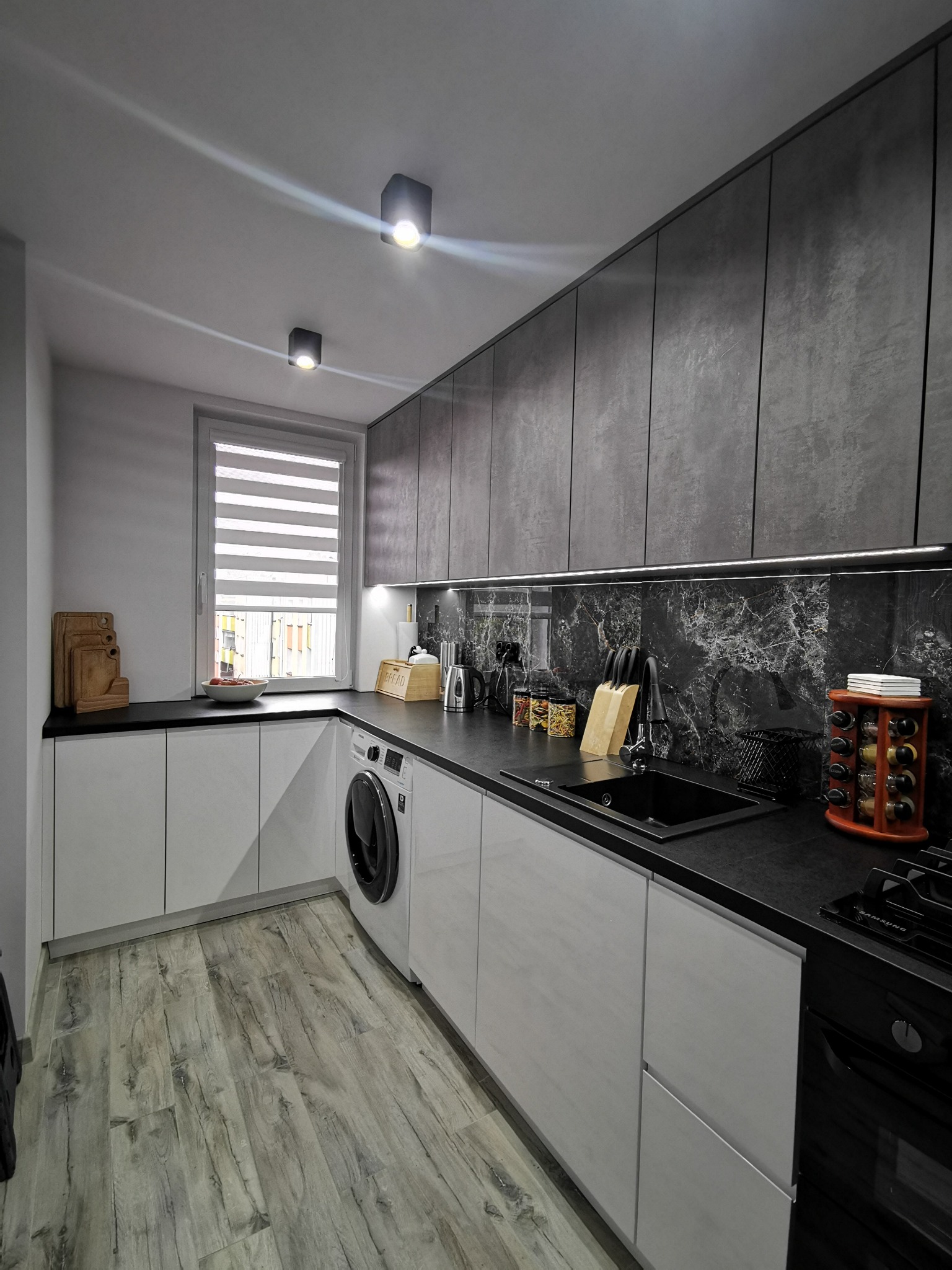 Nowoczesna kuchnia z szarymi górnymi szafkami o teksturze betonu, białymi dolnymi szafkami, czarnym blatem i marmurowym backsplashiem, z pralką w zabudowie.