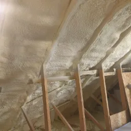 Poddasze z drewnianą konstrukcją pokryte grubą warstwą białej pianki izolacyjnej, widoczne detale faktury pianki i mocowania belek.