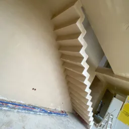 Klatka schodowa po nałożeniu tynku gipsowego, widoczne niezakryte rury instalacyjne przy podłodze i drabina oparta o ścianę w tle.