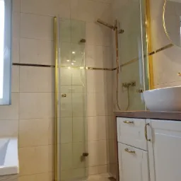 Nowoczesna łazienka z kabiną prysznicową ze złotymi detalami, beżowe płytki na ścianach i biała szafka pod umywalką z złotymi uchwytami.