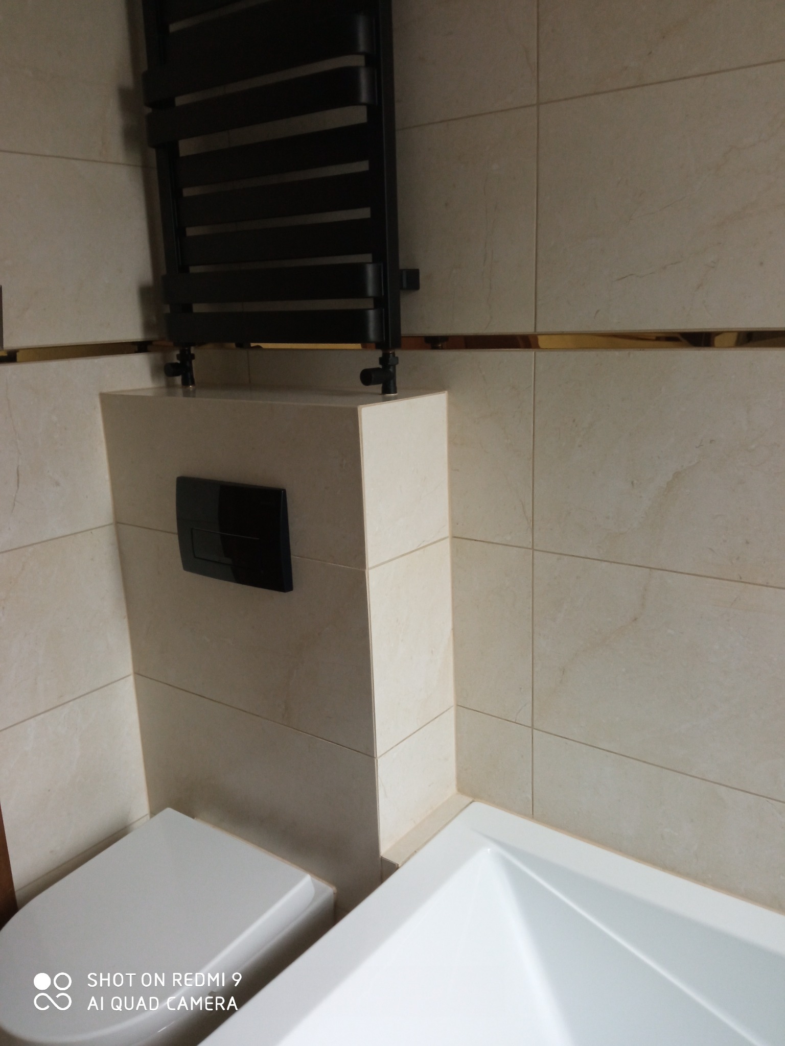 Wykończona łazienka z beżowymi płytkami, czarnym grzejnikiem drabinkowym i przyciskiem spłuczki, widoczny fragment toalety i wanny.