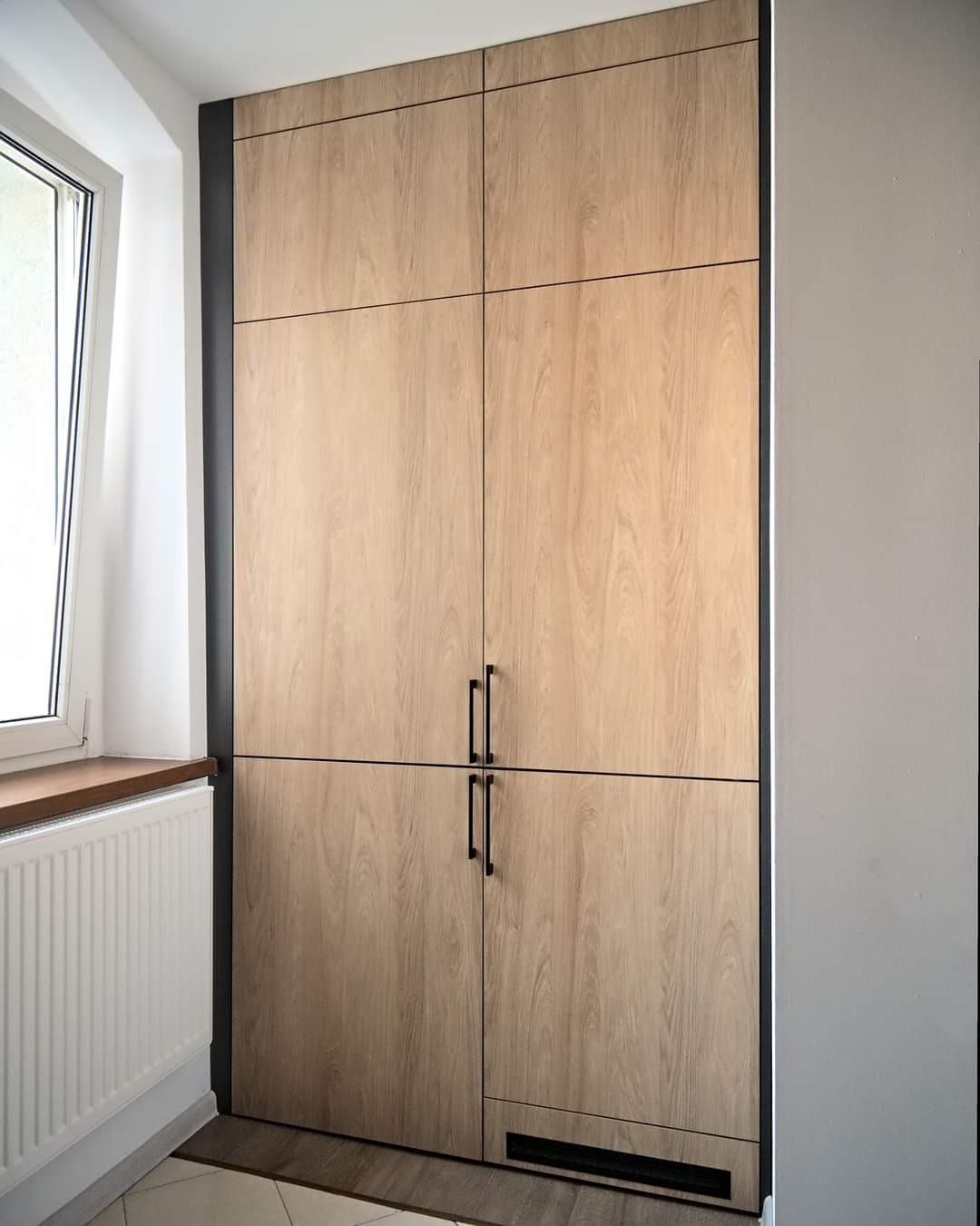 Wysoka, zabudowana szafa z czterema drzwiami o jasnym, drewnopodobnym wykończeniu, z czarnymi uchwytami i obramowaniem, umieszczona we wnęce obok okna i grzejnika.