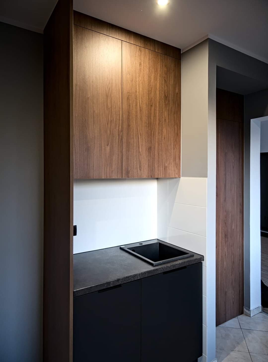 Nowoczesna aranżacja wnętrza z zabudową kuchenną: górne szafki z forniru orzechowego, dolne szafki w kolorze grafitowym z czarnym, minimalistycznym zlewem, białe płytki na ścianie, oraz drzwi...
