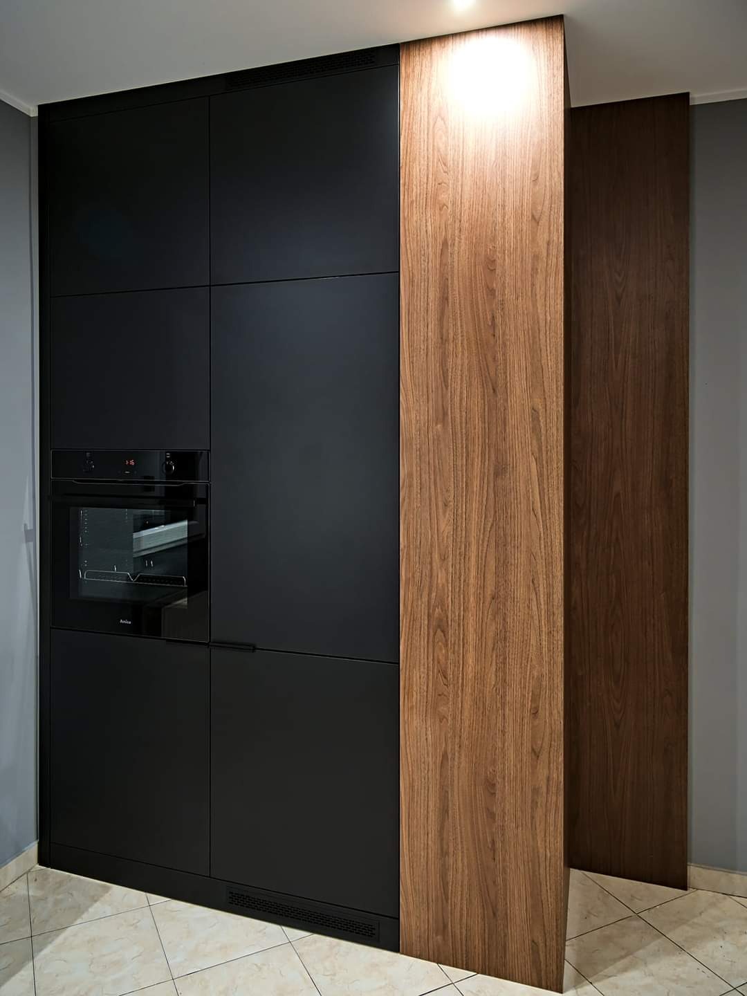 Nowoczesna zabudowa kuchenna: czarne, matowe fronty szafek z wbudowanym piekarnikiem, sąsiadujące z pionowym panelem z drewna orzechowego o wyraźnym usłojeniu.
