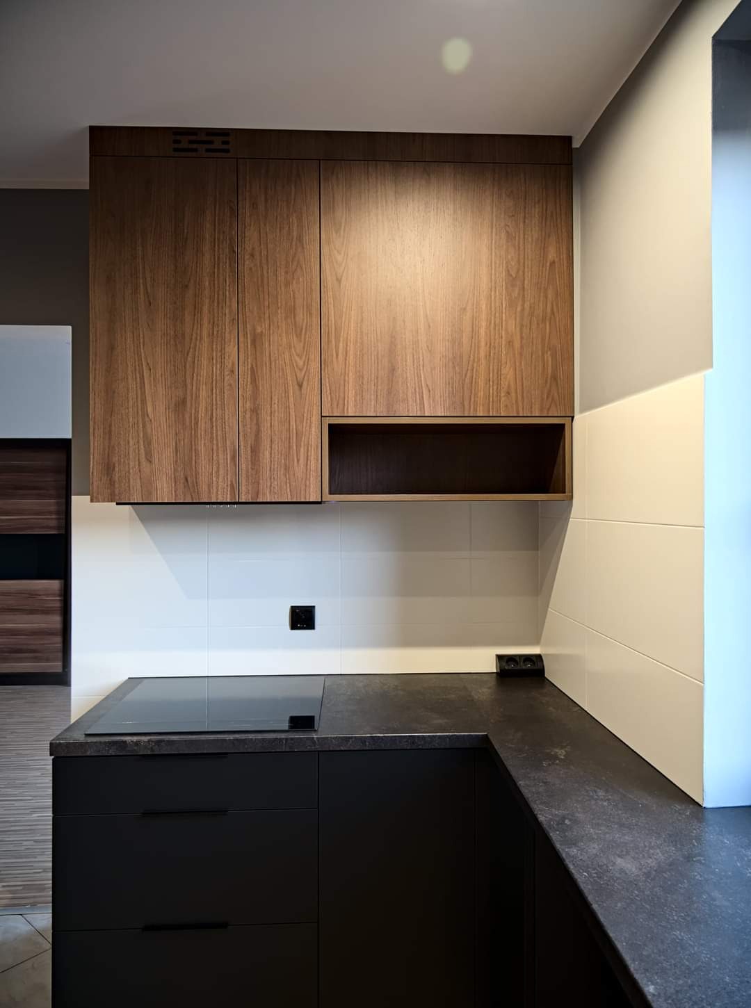 Nowoczesna kuchnia z ciemnymi, matowymi szafkami dolnymi i drewnianymi szafkami górnymi, czarny blat i białe płytki na ścianie, widoczna płyta indukcyjna.