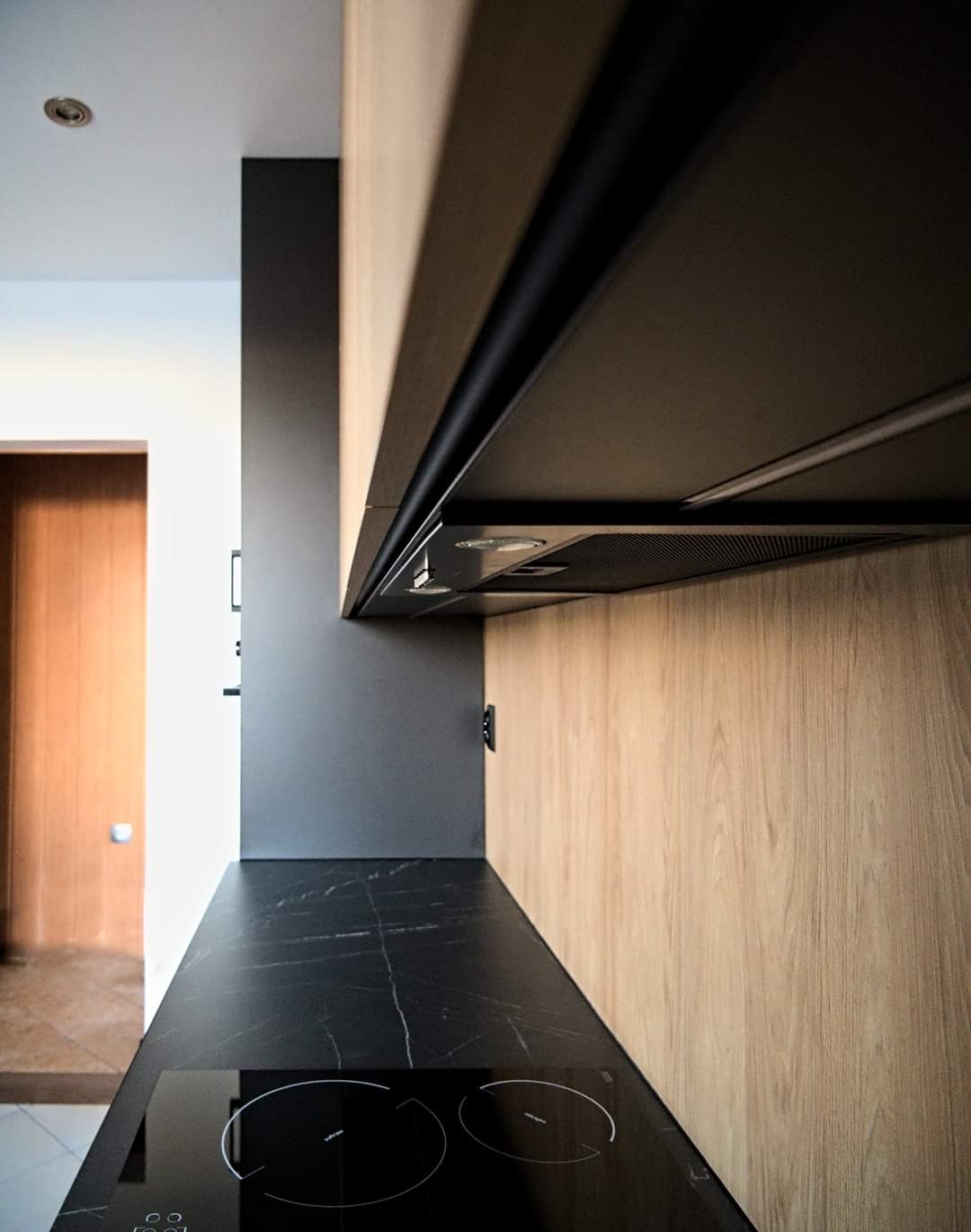 Fragment nowoczesnej kuchni z czarnym blatem imitującym marmur, jasnobrązowymi frontami szafek z wyraźnym usłojeniem drewna i czarnym okapem. Widoczna płyta indukcyjna i fragment ściany pomalowanej...