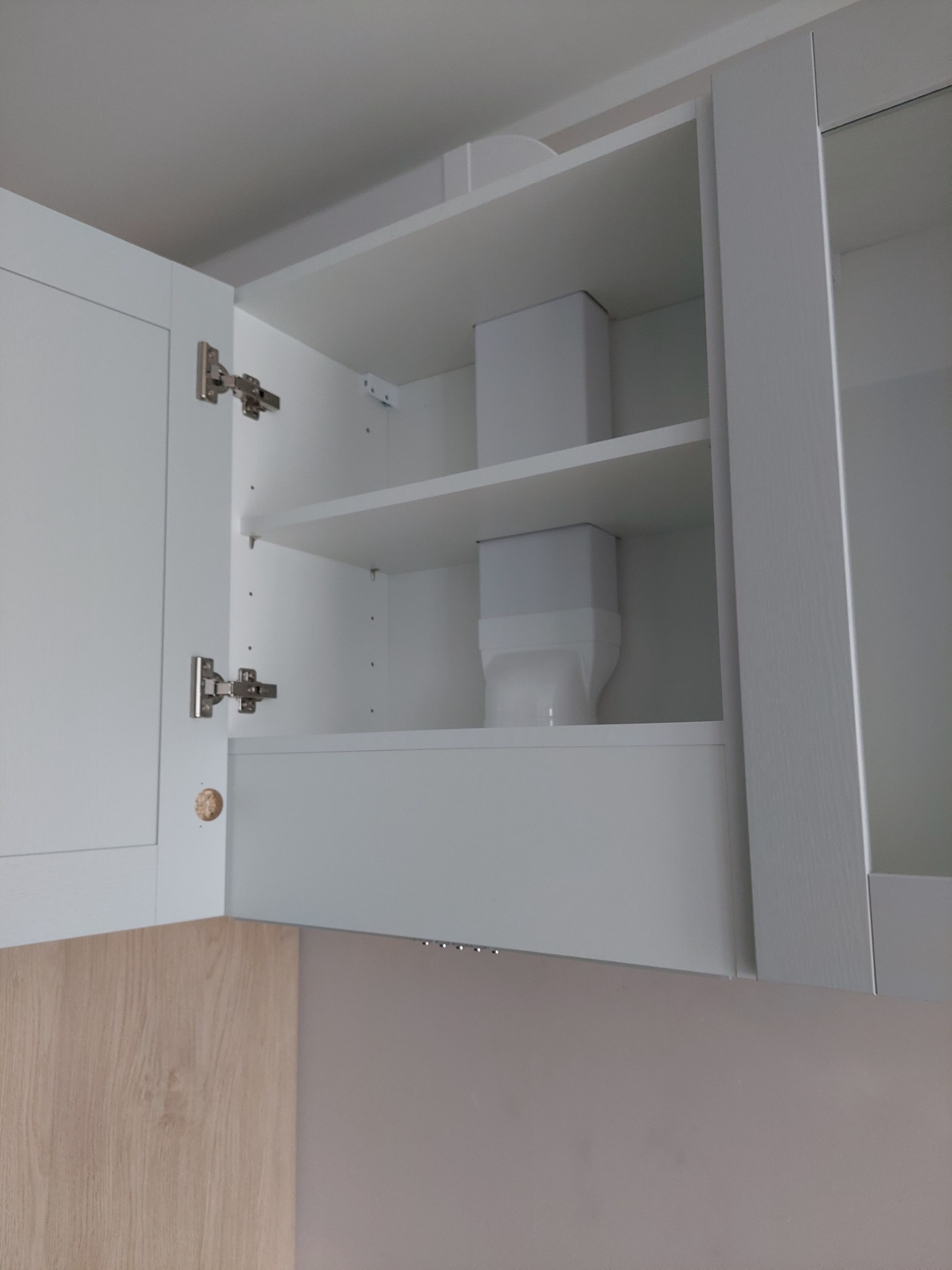 Wnętrze szafki kuchennej z białymi półkami, widoczne zawiasy i fragment frontu szafki w kolorze jasnego drewna.