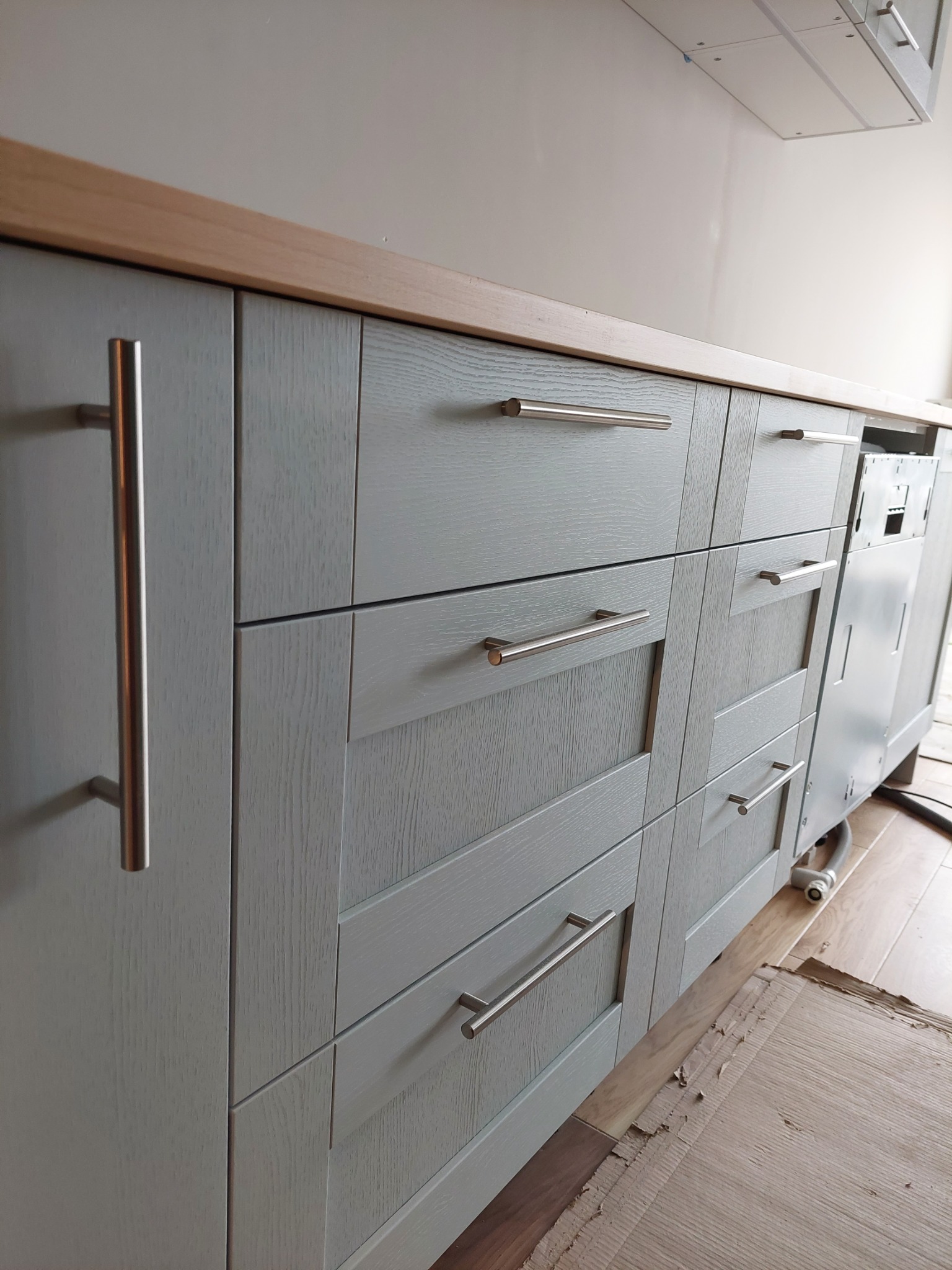 Szare, drewniane szafki kuchenne z długimi, metalowymi uchwytami, widoczna instalacja zmywarki, zabezpieczona podłoga kartonem.