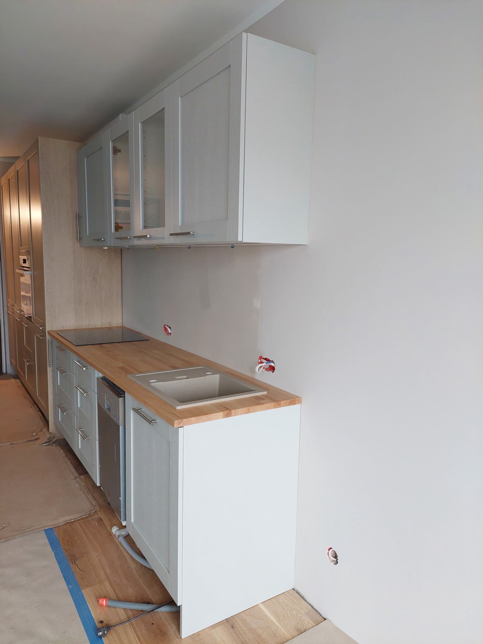 Jasna kuchnia w trakcie montażu: widoczne wiszące szafki z przeszkleniami, drewniany blat, płyta indukcyjna i zlew, instalacje elektryczne i hydrauliczne jeszcze nieukończone, podłoga zabezpieczona...
