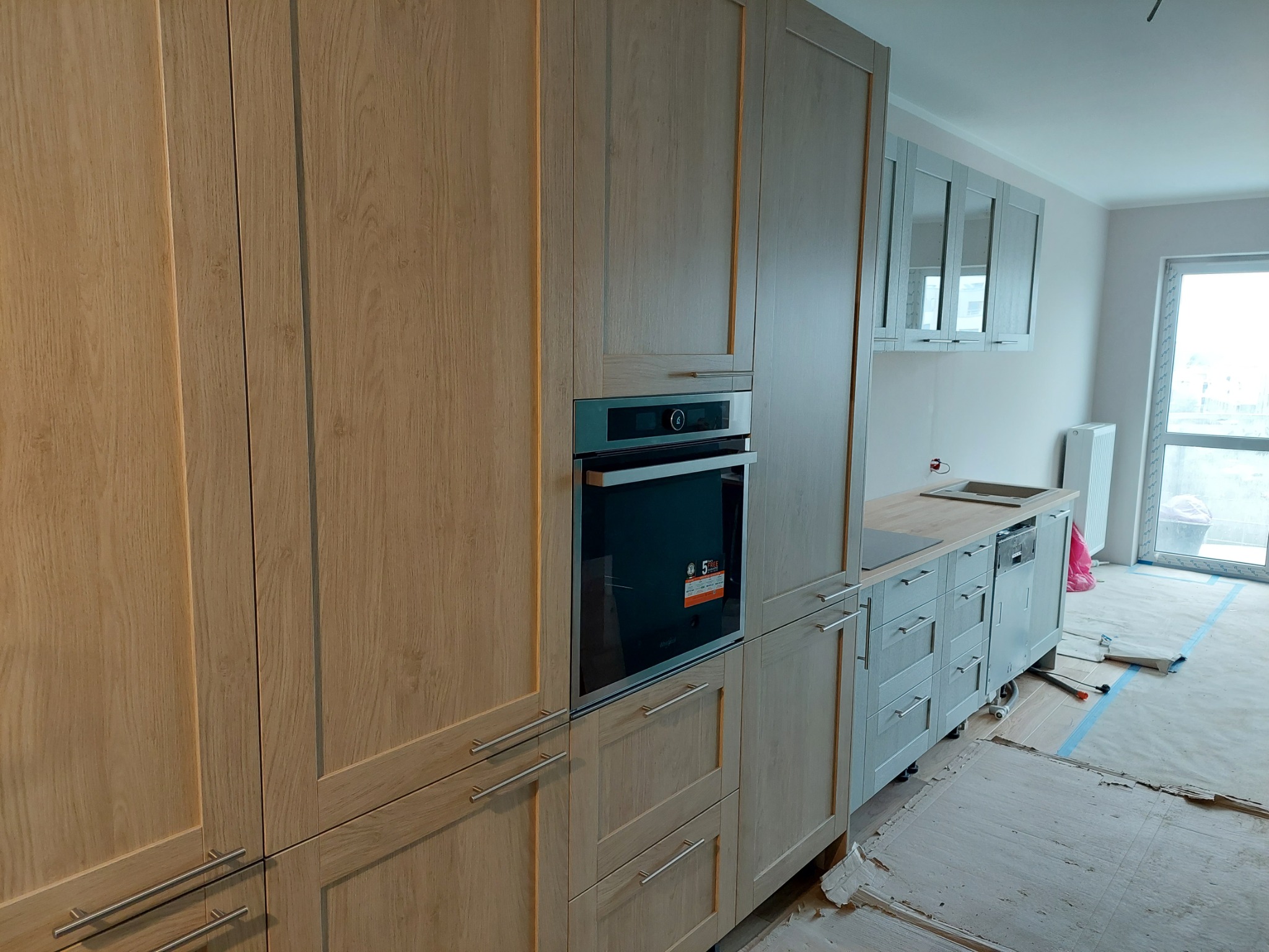 Jasne drewniane szafki kuchenne na wymiar z wbudowanym piekarnikiem, szare szafki wiszące z przeszklonymi frontami, blat z jasnego drewna, podłoga zabezpieczona kartonem.