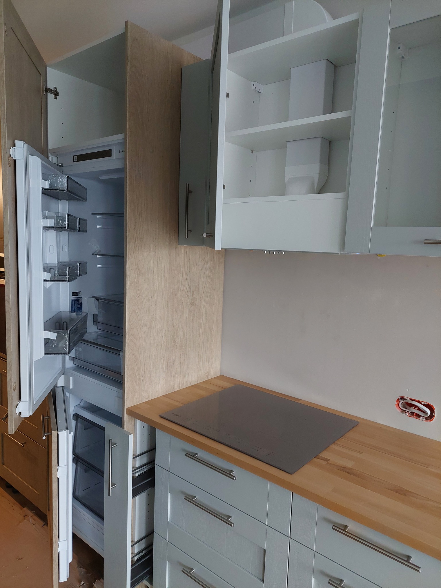 Jasna kuchnia z drewnianym blatem, wbudowaną płytą indukcyjną i lodówką z otwartymi drzwiami, widoczne wnętrze szafek wiszących i szuflad z metalowymi uchwytami.