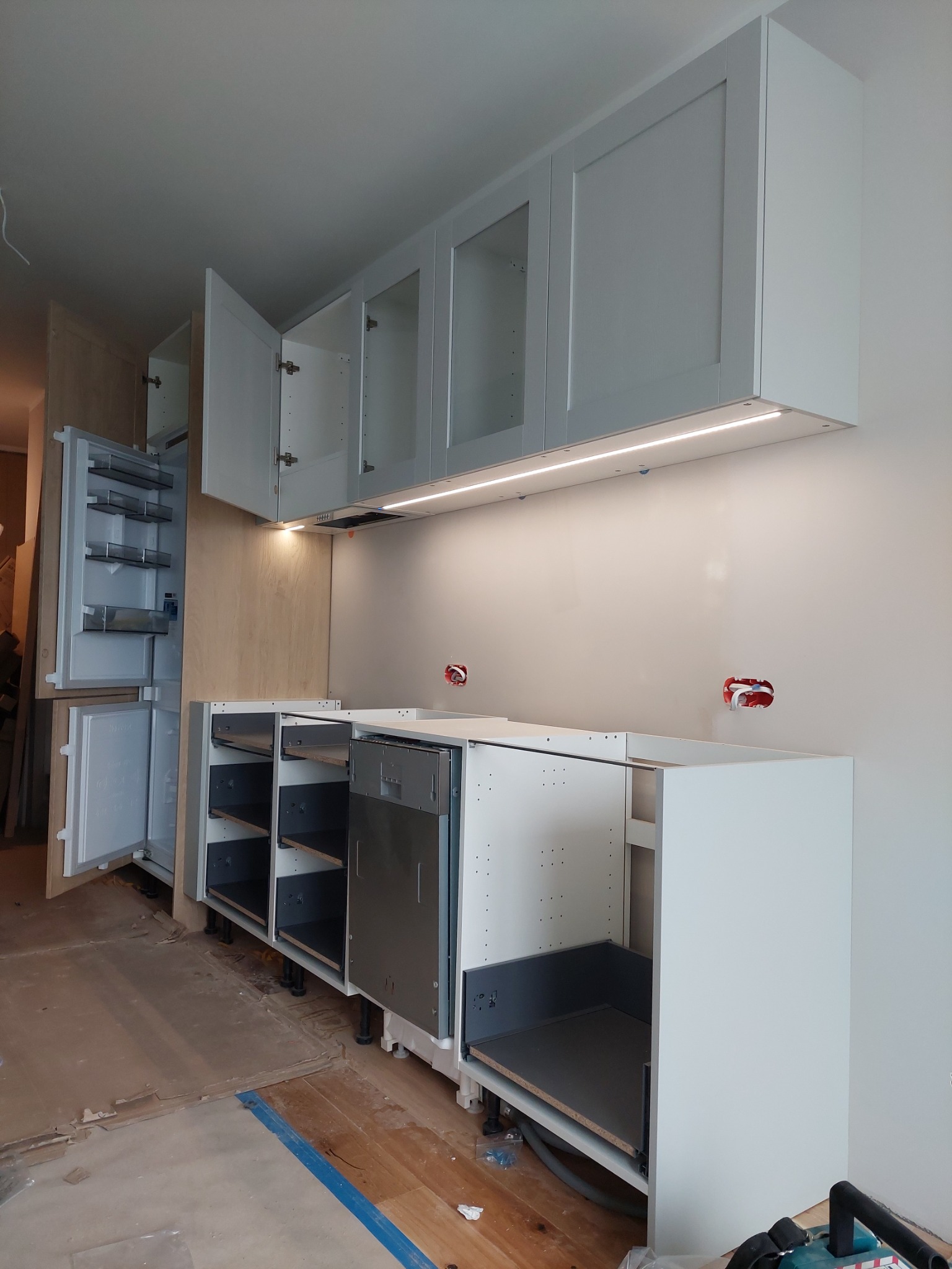 Montaż białej zabudowy kuchennej: widoczne szafki górne z oświetleniem LED, dolne szafki z szufladami i frontami, oraz otwarta lodówka w zabudowie, na podłodze widoczne ślady po remoncie.