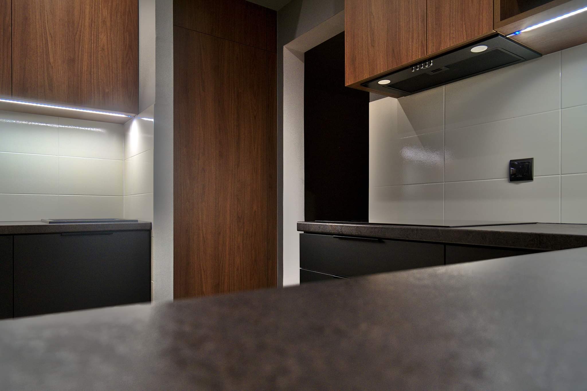 Nowoczesna kuchnia z ciemnymi szafkami dolnymi i drewnianymi górnymi, podświetlona listwą LED, z widocznym fragmentem blatu i okapem.