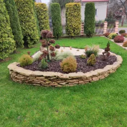 Owalna rabata kwiatowa otoczona kamiennym murkiem z piaskowca, wypełniona korą, białym żwirem i różnorodną roślinnością ozdobną, na tle zadbanego trawnika, tuj i ogrodzenia.