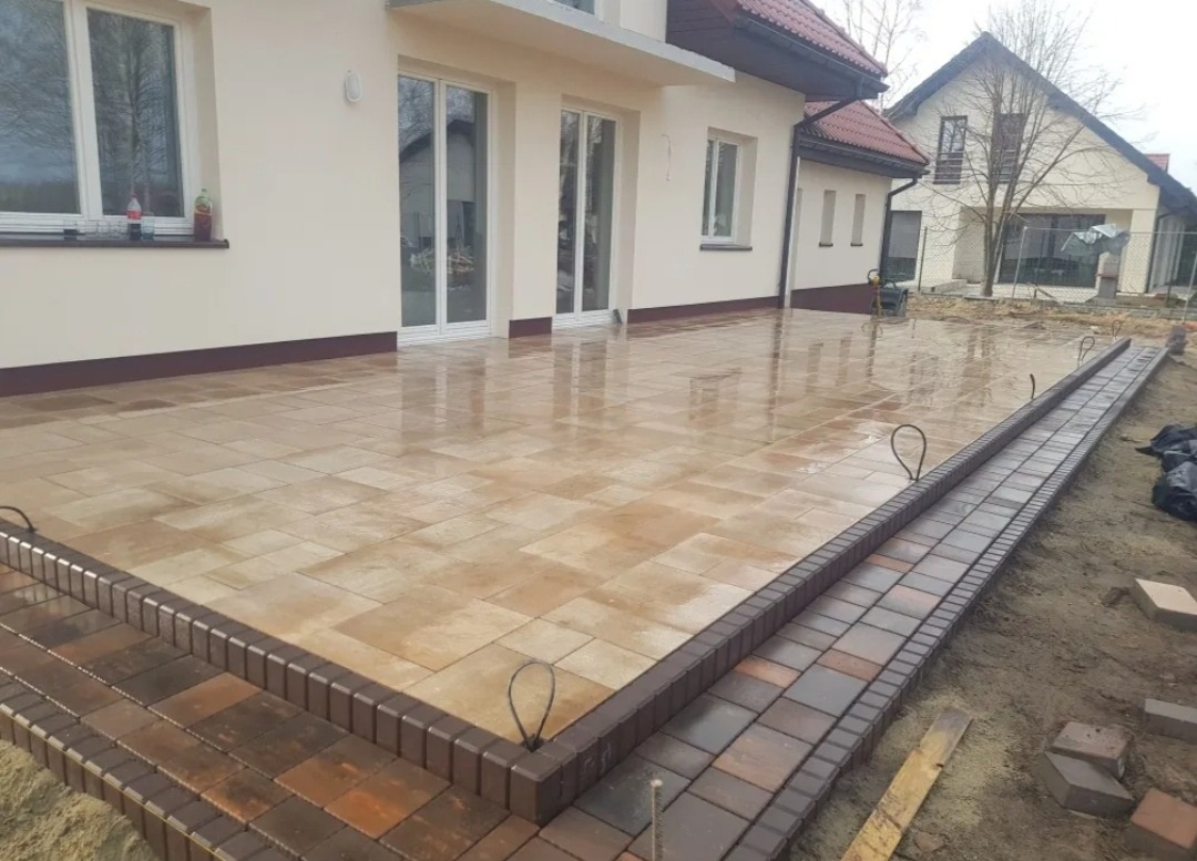 Świeżo ułożona, mokra kostka brukowa w odcieniach beżu i brązu tworząca taras przed domem. Widoczne obrzeża z ciemniejszej kostki i przygotowane podłoże pod dalszą część chodnika.