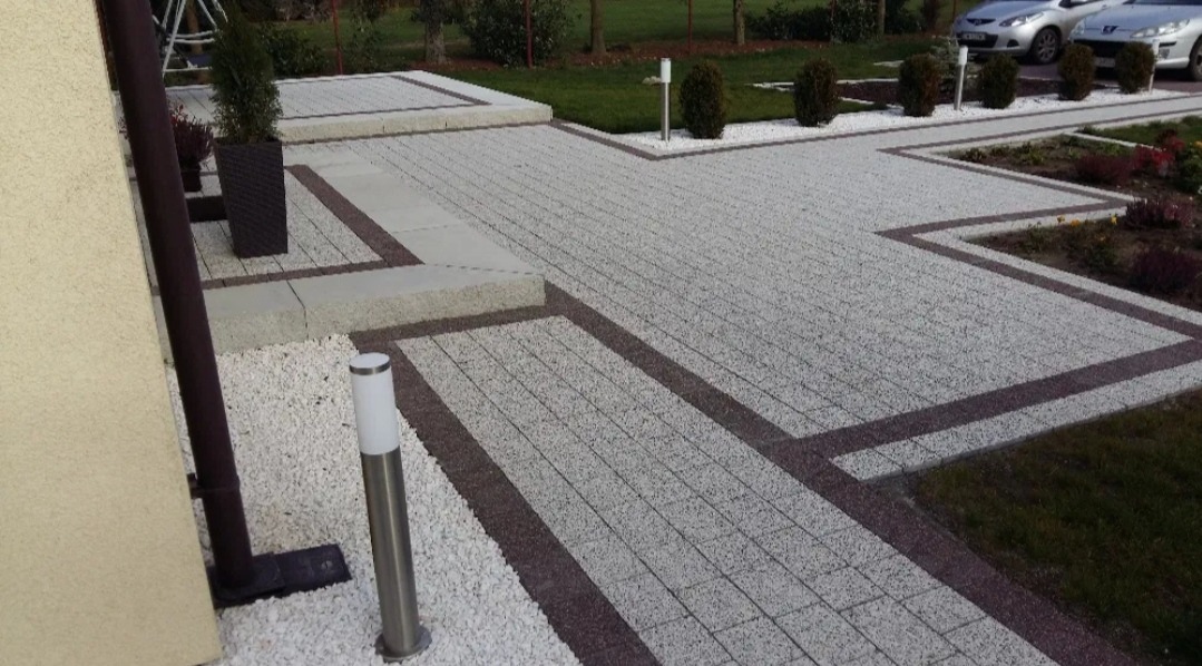Ułożona jasnoszara kostka brukowa z ciemniejszym obramowaniem, tworząca ścieżkę i podjazd przed domem, z białym kamieniem ozdobnym, lampami ogrodowymi i zielonym trawnikiem.