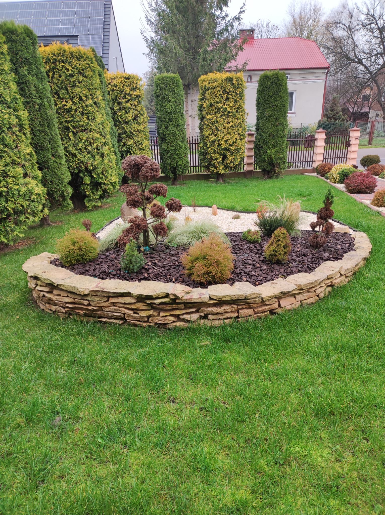 Owalna rabata kwiatowa otoczona kamiennym murkiem z piaskowca, wypełniona korą, białym żwirem i różnorodną roślinnością ozdobną, na tle zadbanego trawnika, tuj i ogrodzenia.