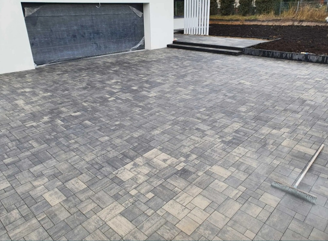 Ułożona kostka brukowa w odcieniach szarości i beżu na podjeździe przed domem z garażem, widoczna szczotka do zamiatania.