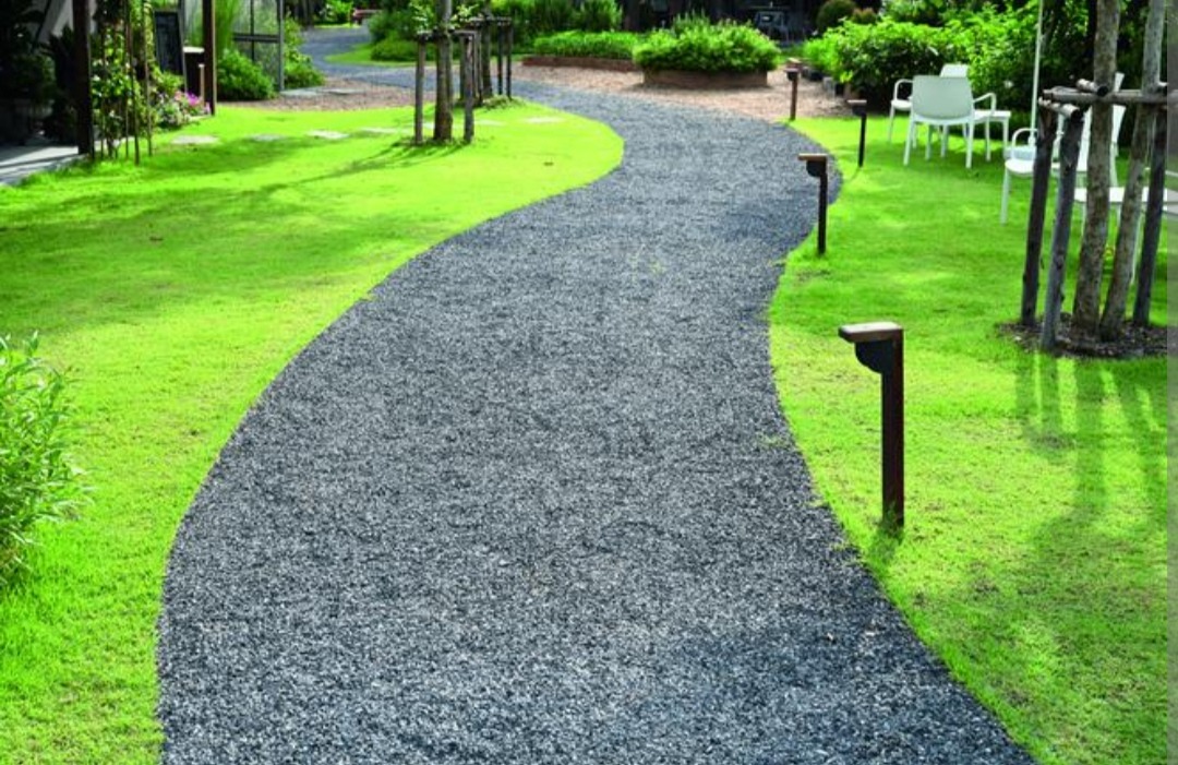 Kręta ścieżka z drobnego, szarego żwiru, otoczona intensywnie zielonym trawnikiem i oświetlona niskimi lampami ogrodowymi, prowadząca do białych krzeseł i stołu w oddali.