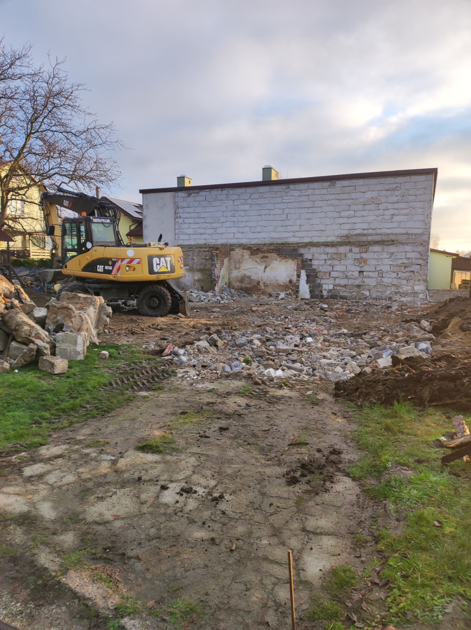 Żółta koparka CAT M318D na placu budowy z gruzem i fragmentami ściany z białej cegły