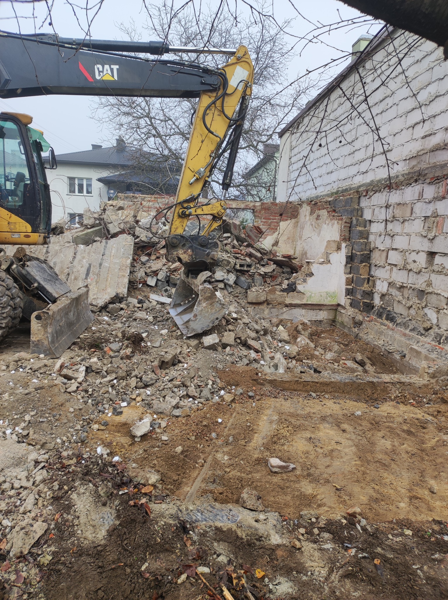 Żółta koparka CAT podczas rozbiórki starego budynku z cegły i betonu, gruzowisko na placu budowy, widoczne fragmenty ściany i fundamentów.