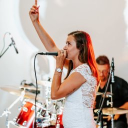 Zespół z muzyką na żywo ,STAND-ART, - Wokalistka w białej sukience śpiewa do mikrofonu, w tle perkusista. Kobieta ma uniesioną rękę, na nadgarstku zegarek. Scena oświetlona ciepłym światłem.