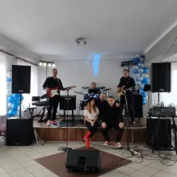 Zespół na scenie podczas występu w sali z dekoracjami balonowymi, wokalistka w czerwonych kozakach siedzi na skraju sceny, a w tle widoczny perkusista.