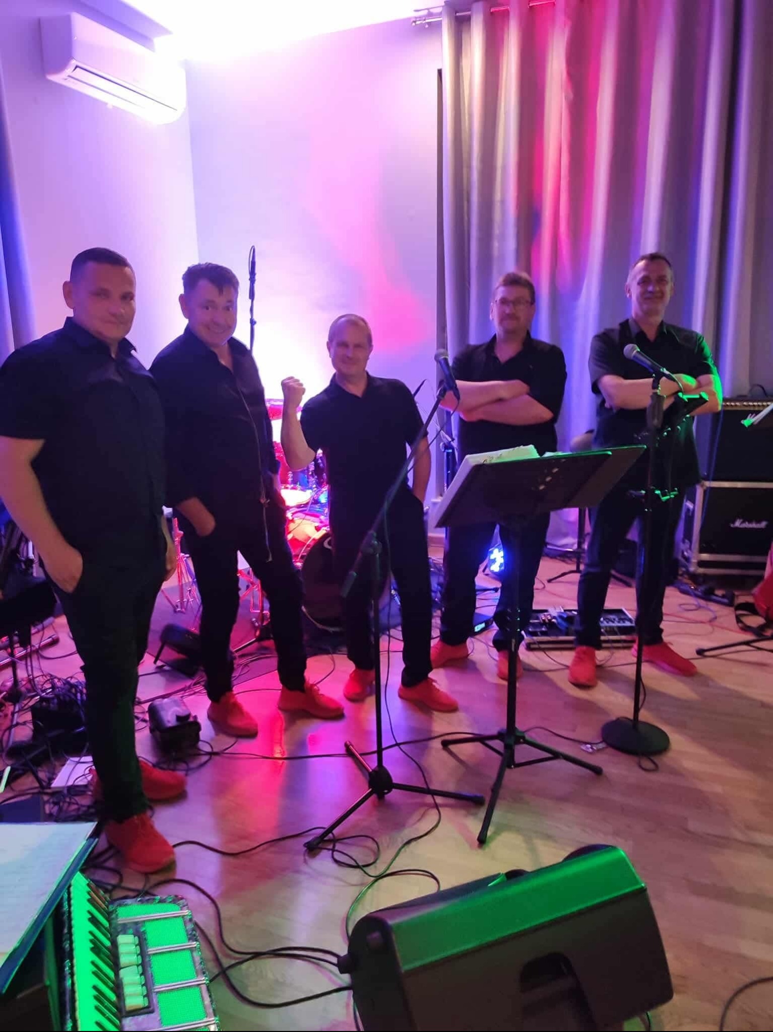 Zespół muzyczny w czarnych strojach i czerwonych butach pozuje na scenie z instrumentami, oświetlony kolorowymi światłami. Widoczny akordeon i perkusja.