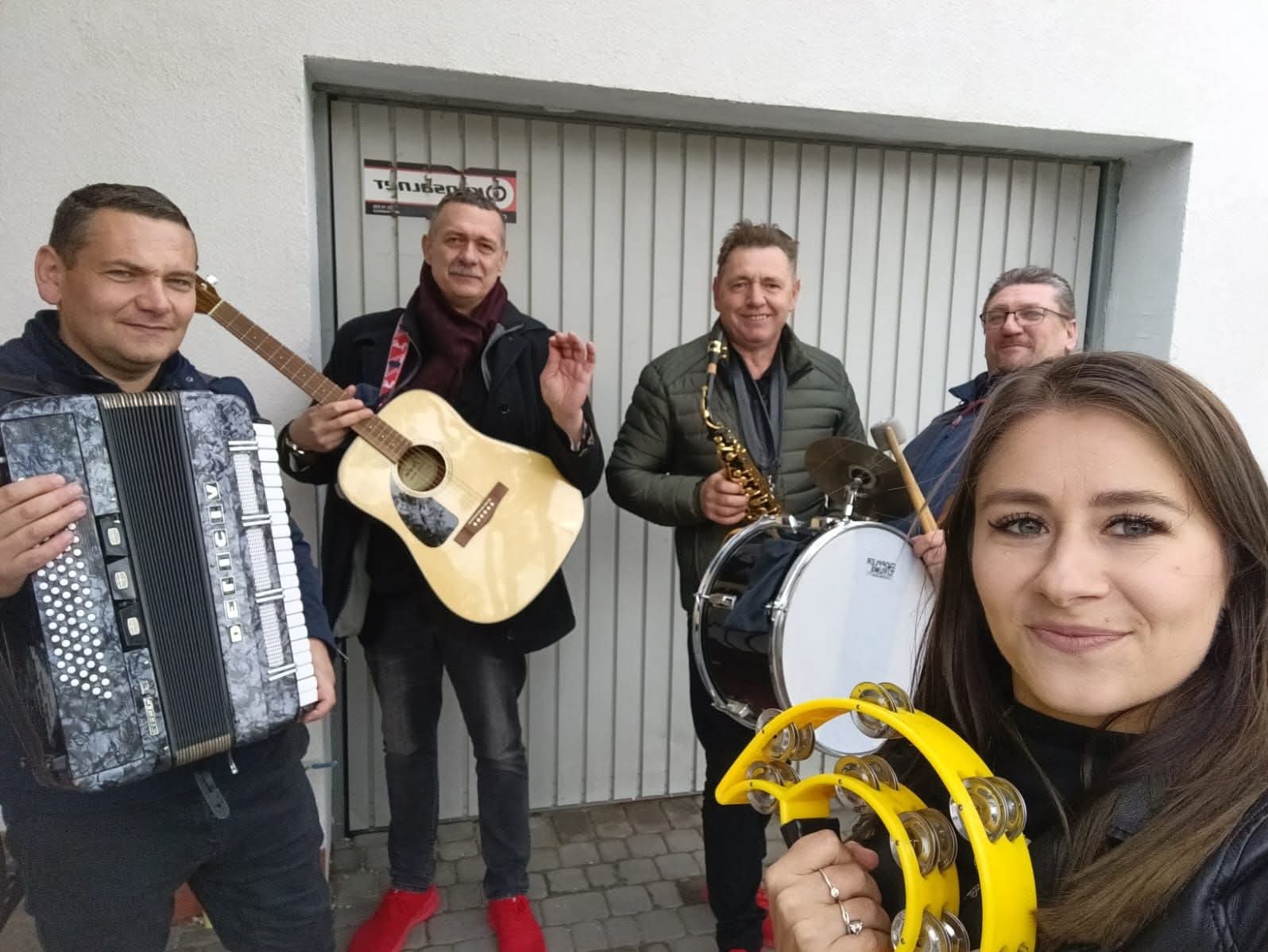 Zespół muzyczny w plenerze: akordeon, gitara, saksofon, perkusja i tamburyn. Uśmiechnięci muzycy gotowi do występu, z przodu kobieta z tamburynem.