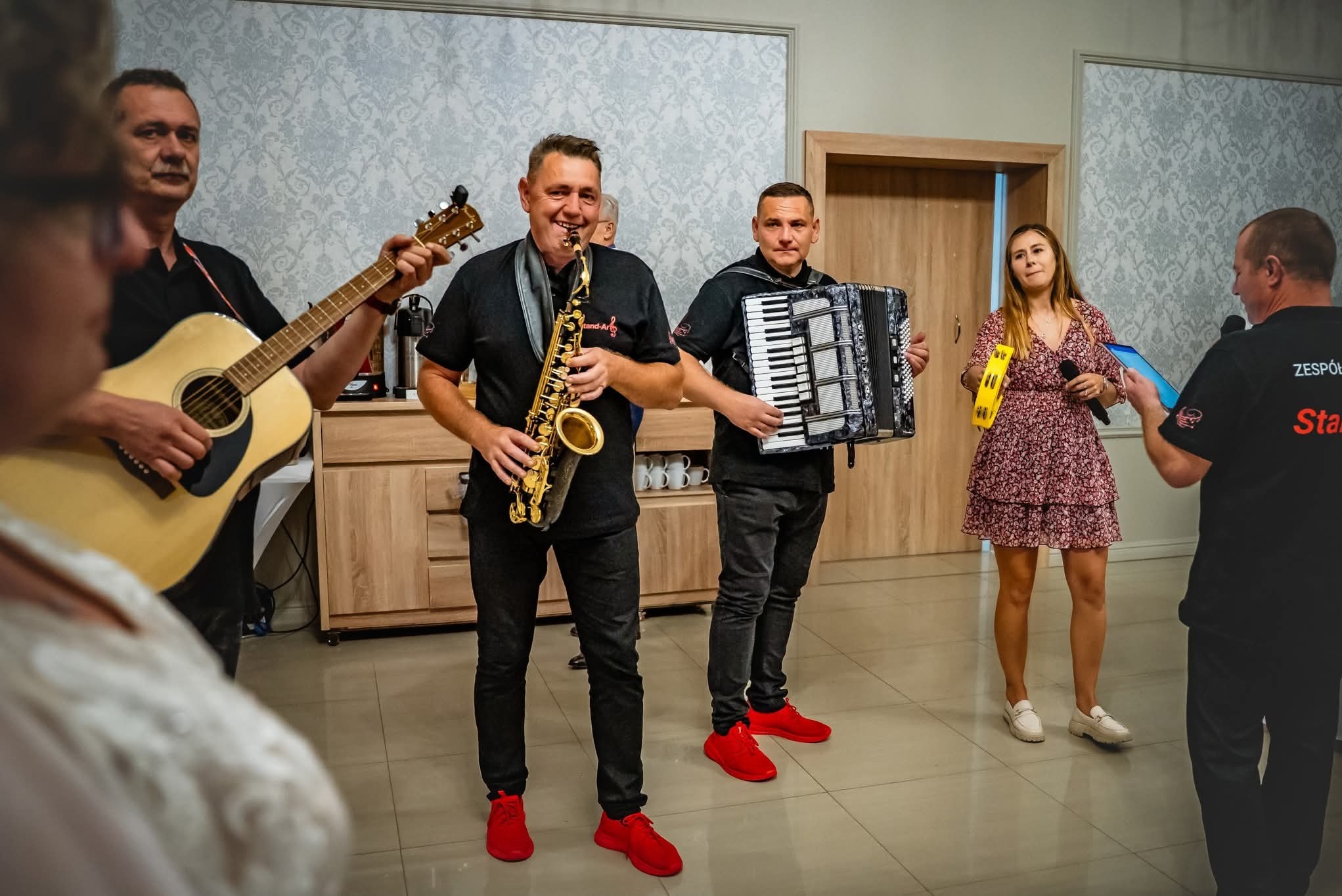 Zespół muzyczny w trakcie występu: saksofonista z uśmiechem, akordeonista, gitarzysta i wokalistka z tamburynem. Wszyscy w czarnych koszulkach i czerwonych butach.