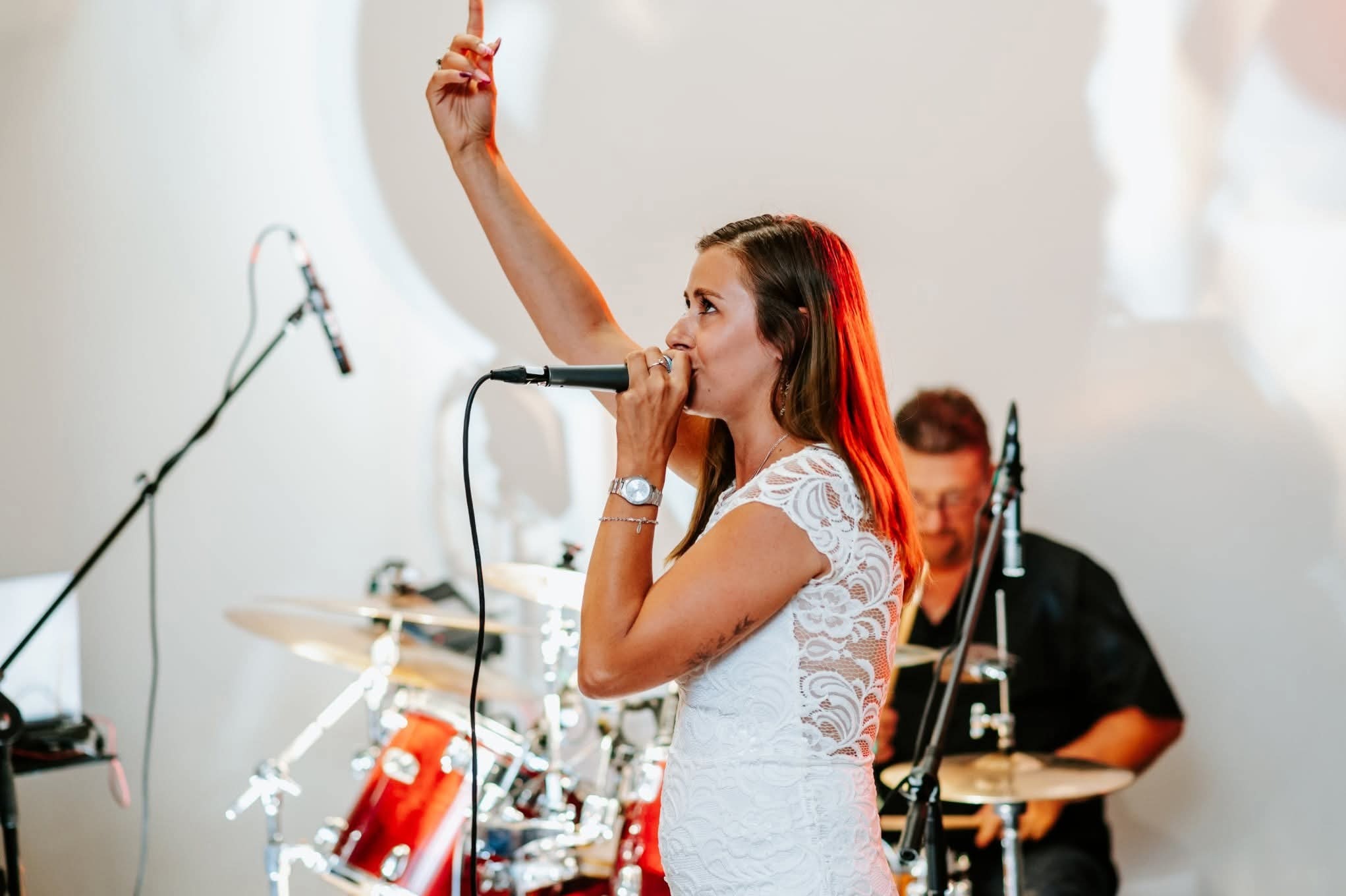 Wokalistka w białej sukience śpiewa do mikrofonu, w tle perkusista. Kobieta ma uniesioną rękę, na nadgarstku zegarek. Scena oświetlona ciepłym światłem.