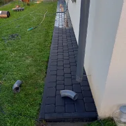 Ciemnoszara kostka brukowa ułożona wzdłuż ściany domu z rurą spustową, widoczne złącza drenażowe na trawie, w tle rozładowany samochód i narzędzia.