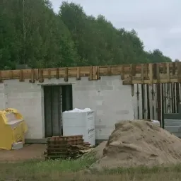 Wznoszone ściany domu z bloczków betonowych, widoczne drewniane szalunki stropu, sterta piasku i materiały budowlane na placu budowy, w tle las.