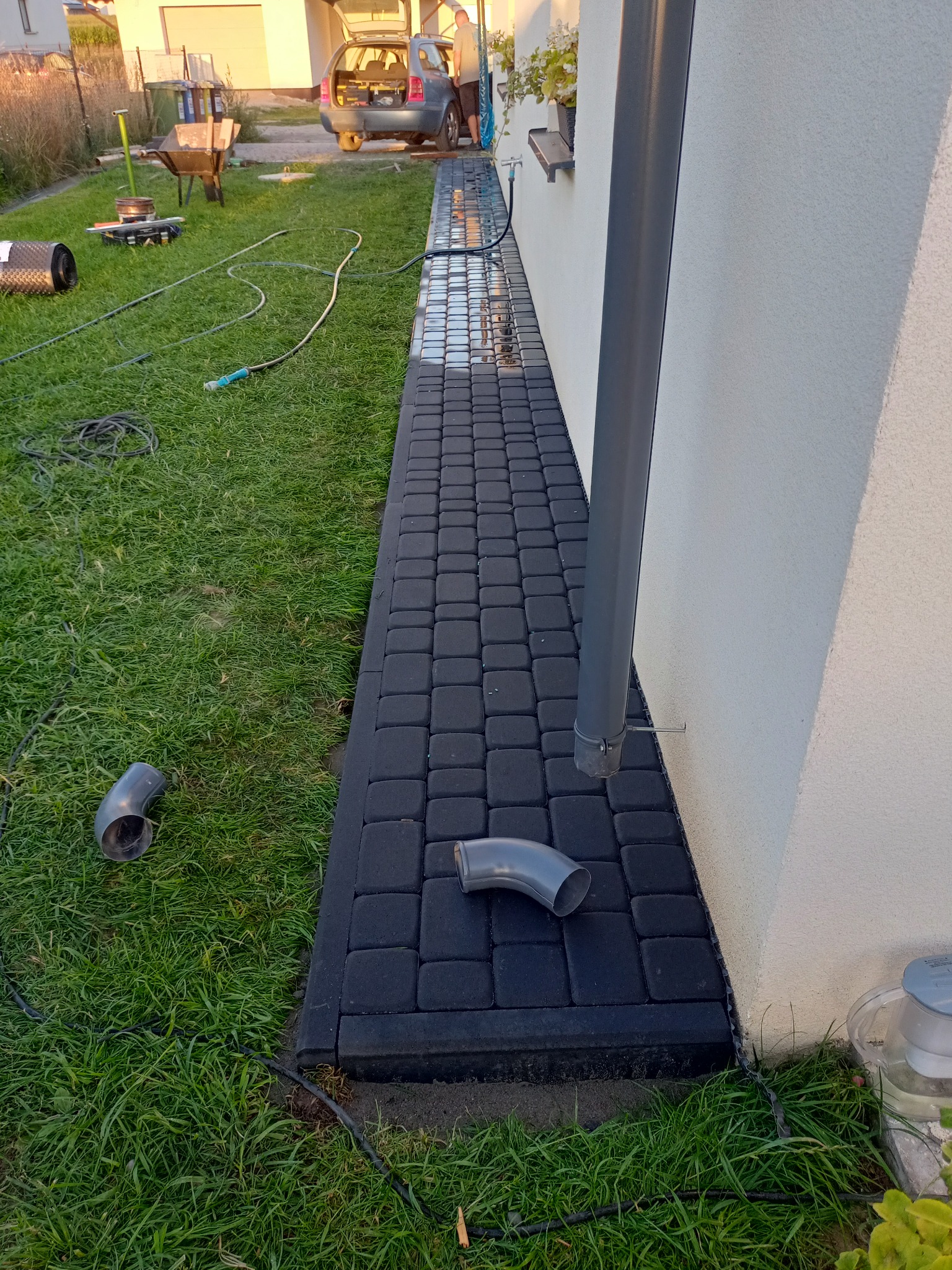 Ciemnoszara kostka brukowa ułożona wzdłuż ściany domu z rurą spustową, widoczne złącza drenażowe na trawie, w tle rozładowany samochód i narzędzia.