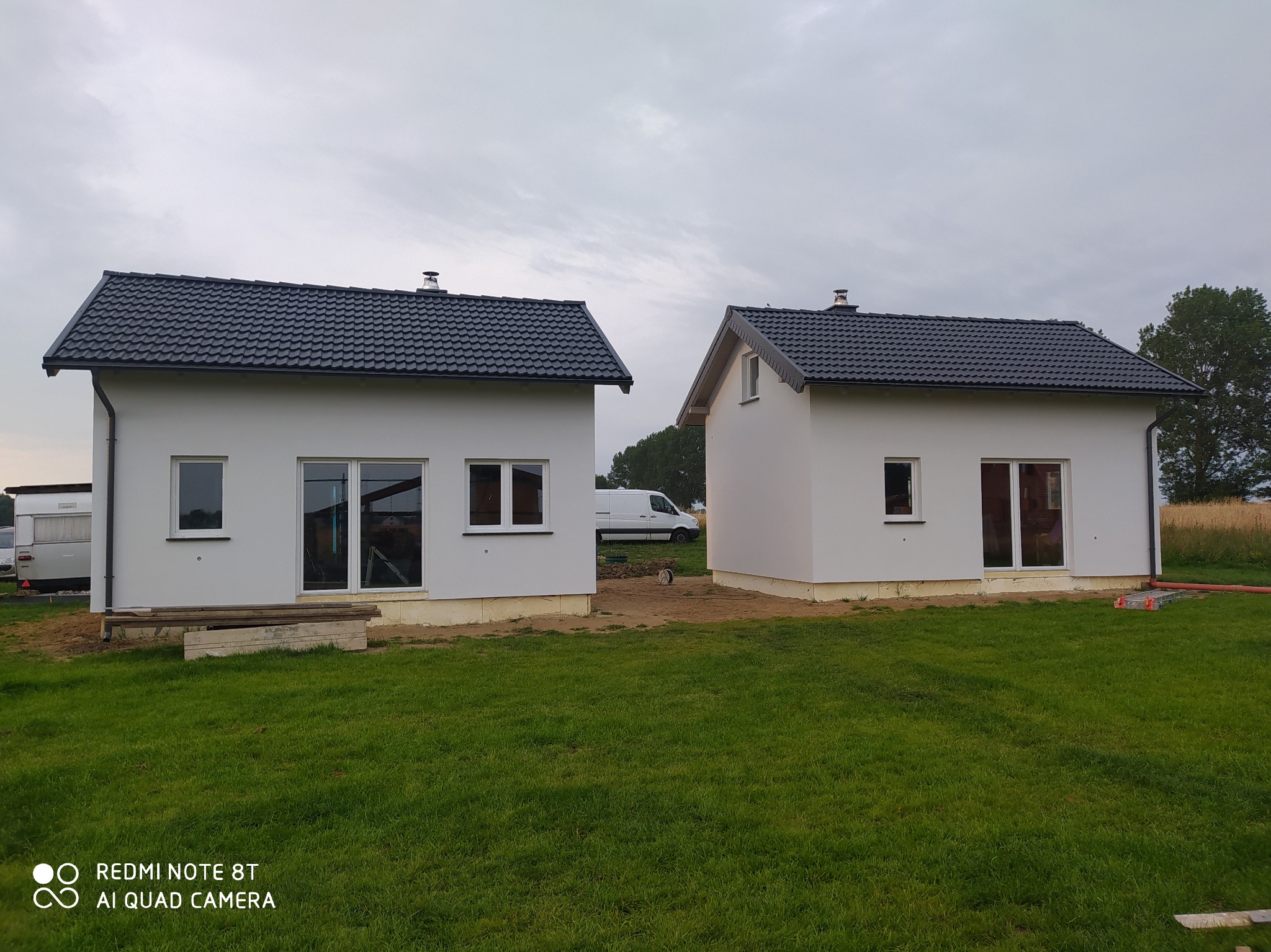 Dwa nowo wybudowane, białe domy jednorodzinne z ciemnoszarą dachówką, widoczne z poziomu trawnika w pochmurny dzień, z zaparkowanym białym vanem w tle.