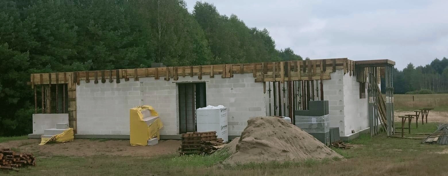 Wznoszone ściany domu z bloczków betonowych, widoczne drewniane szalunki stropu, sterta piasku i materiały budowlane na placu budowy, w tle las.