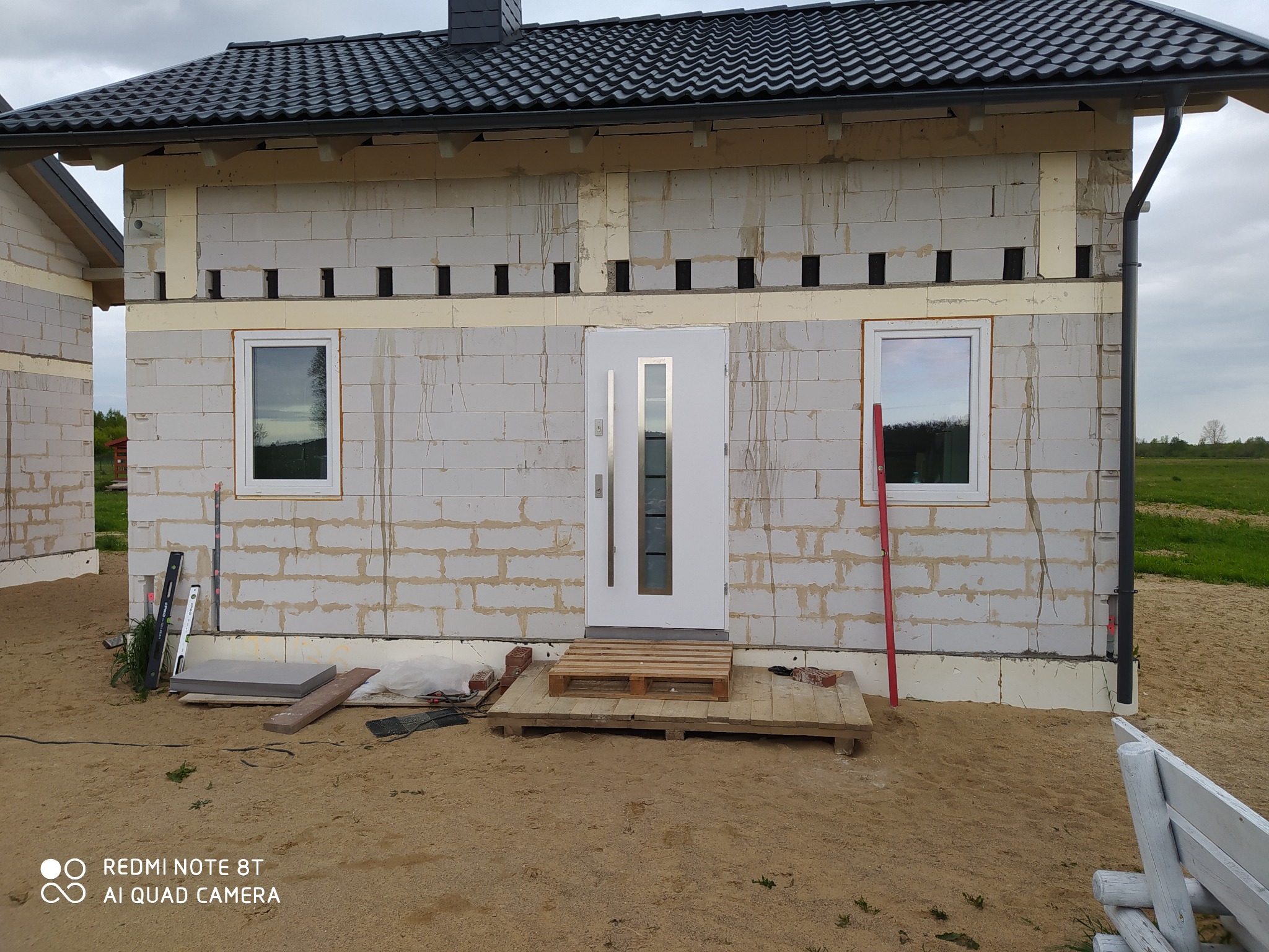 Surowy budynek w trakcie budowy, ściany z białych bloczków, widoczne ocieplenie, tymczasowe wejście z palet, narzędzia budowlane leżące obok, piasek na podwórku.