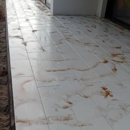 Wielki Stroy - Jasnoszare płytki tarasowe z nieregularnymi, brązowymi wzorami, ułożone wzdłuż nowoczesnego budynku ze szklanymi drzwiami. Widoczna wilgoć na powierzchni i luźna ziemia przy krawędzi.