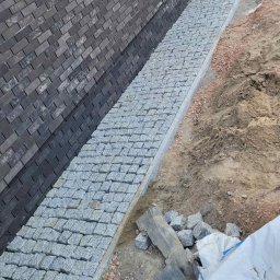 Wielki Stroy - Wąski pas chodnika z jasnoszarej kostki granitowej, ułożony wzdłuż ciemnej ściany z cegły, z widocznym surowym gruntem i resztkami materiałów budowlanych.