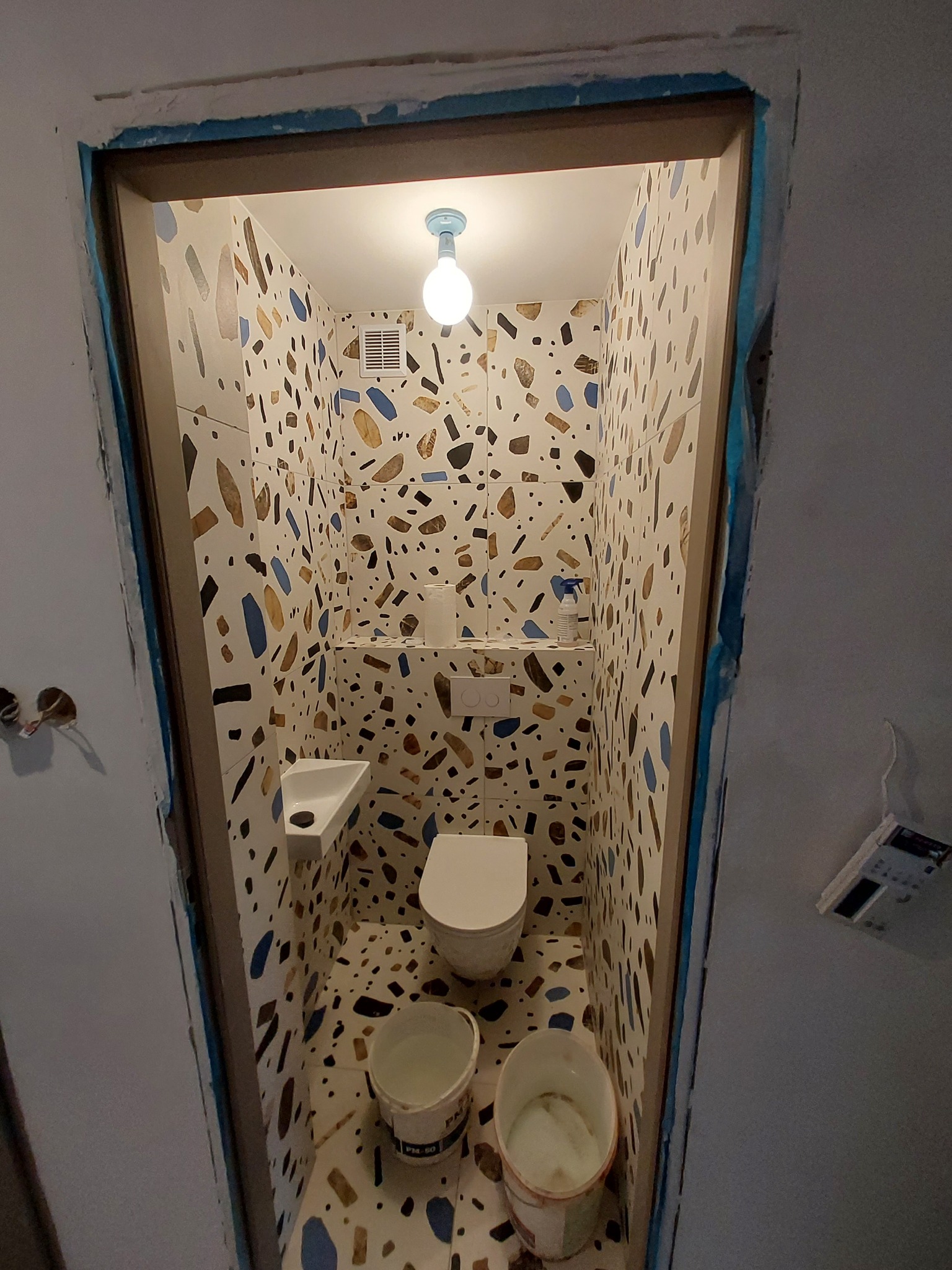 Wąska toaleta w trakcie prac wykończeniowych, ściany i podłoga wyłożone płytkami z motywem lastryko, widoczne dwa wiadra z farbą i zabezpieczone futryny drzwi.