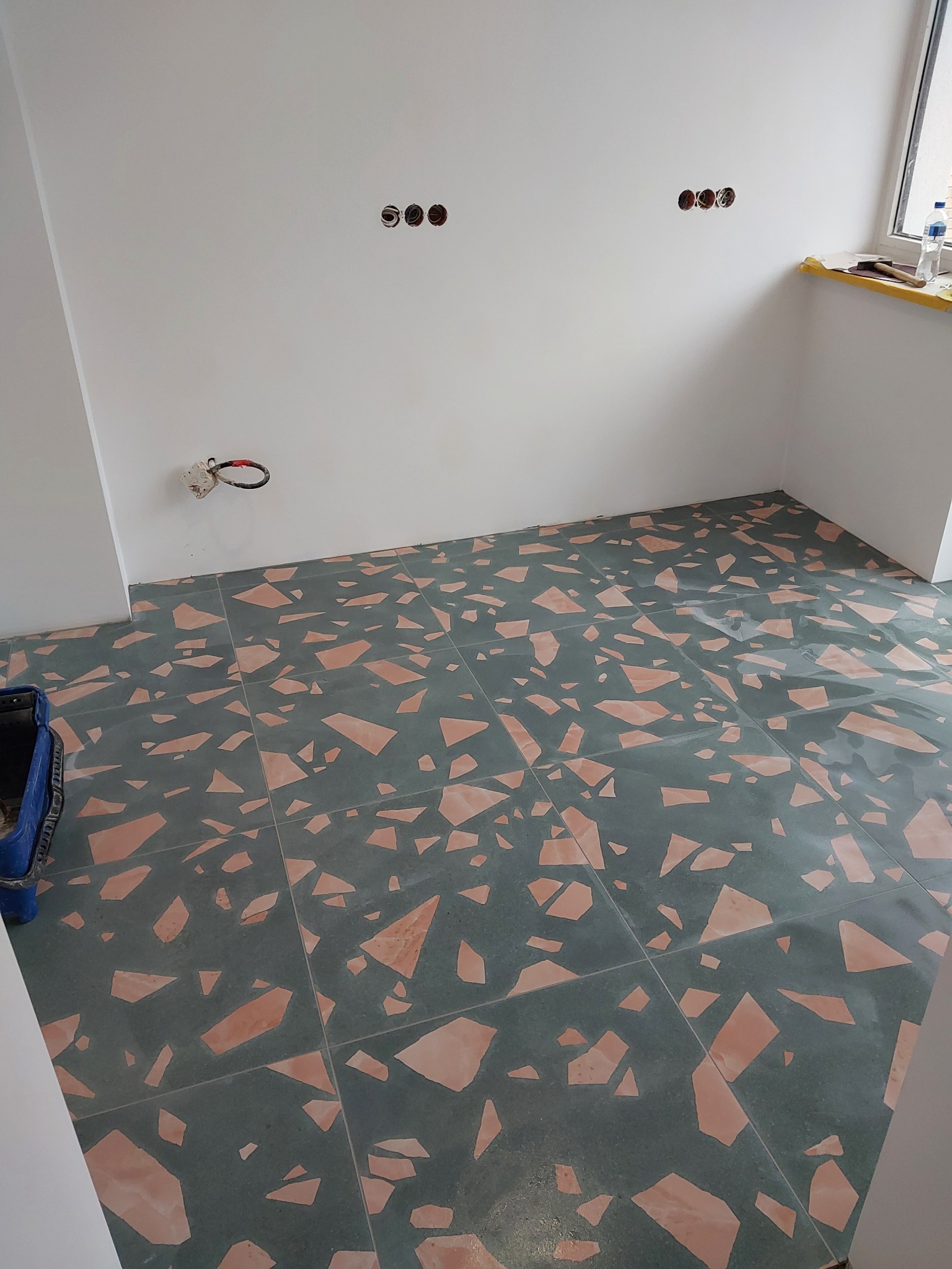 Nowo położona, wzorzysta podłoga z płytek w odcieniach szarości i terakoty w pustym pomieszczeniu z białymi ścianami i przygotowanymi gniazdkami elektrycznymi.